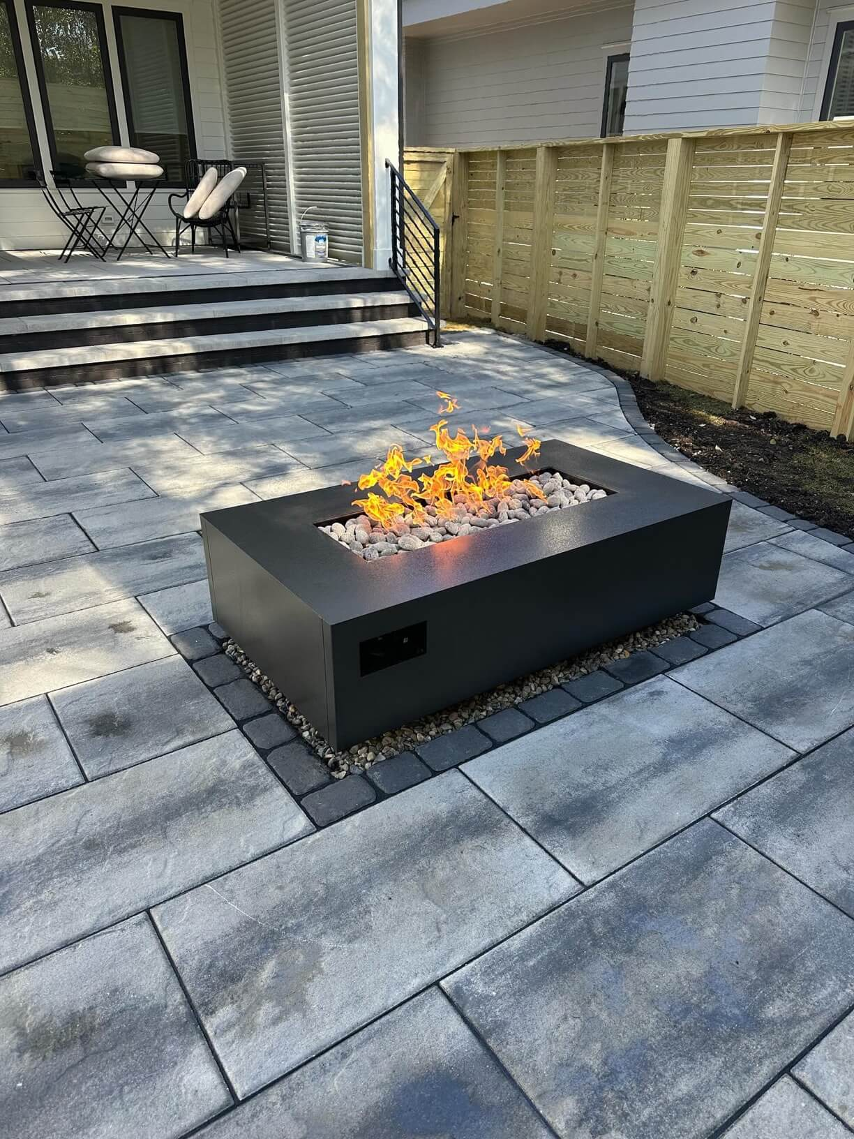 A backyard patio featuring a modern rectangular fire pit with flames, surrounded by large gray stone pavers, and bordered by a wooden fence and a house with a small porch.