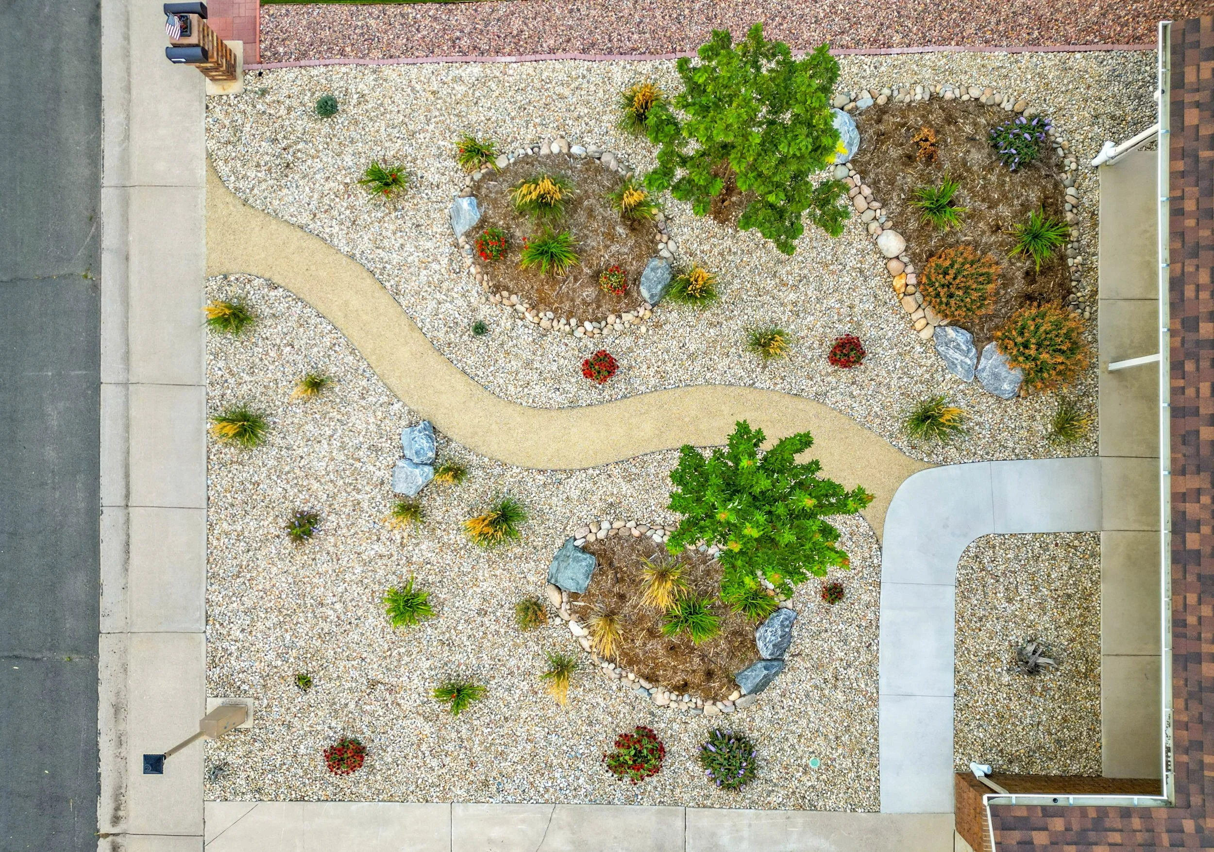A landscaped front yard with a curving gravel path, small trees, bushes, and flower beds surrounded by rocks, with a sidewalk and street along the edges.