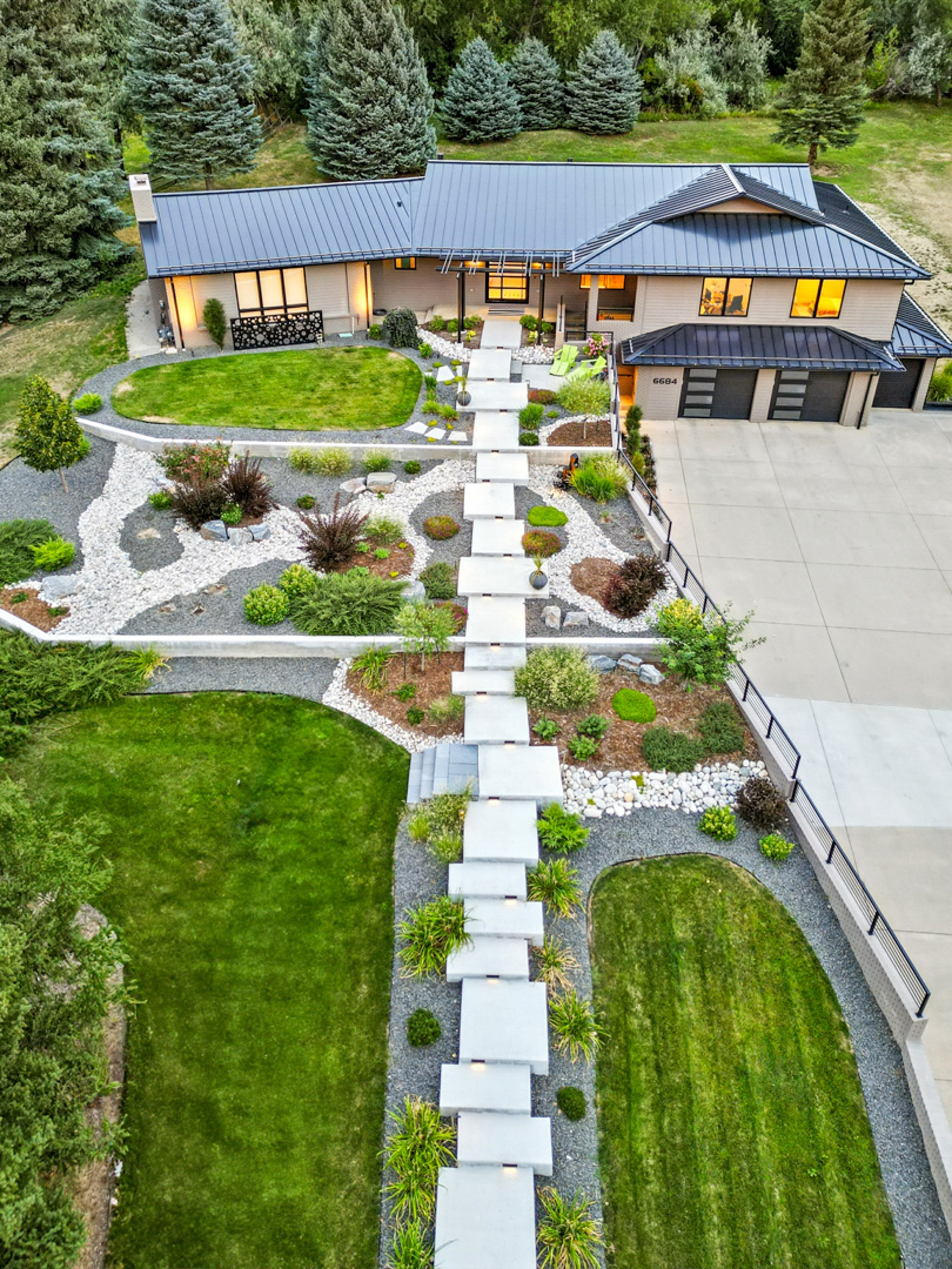 Aerial view of a modern house with a dark metal roof, large windows, and a two-car garage. The front yard features a walkway lined with small plants and lights, a well-maintained lawn, and landscaped garden areas with rocks, shrubs, and small trees. Tall pine trees are visible in the background.