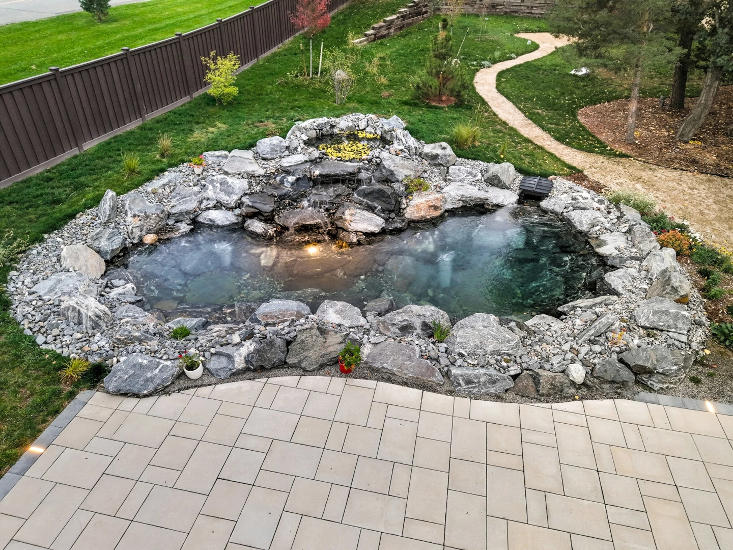 Backyard with a rock-lined pond with flowing water, potted plants, a stone pathway, a grassy lawn, trees, and a brown privacy fence.