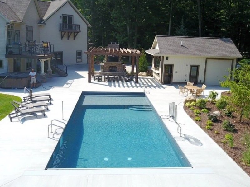 Backyard pool area with a rectangular swimming pool, lounge chairs, a shaded pergola with outdoor fireplace, and a small building with a deck and dining table, surrounded by greenery and trees.