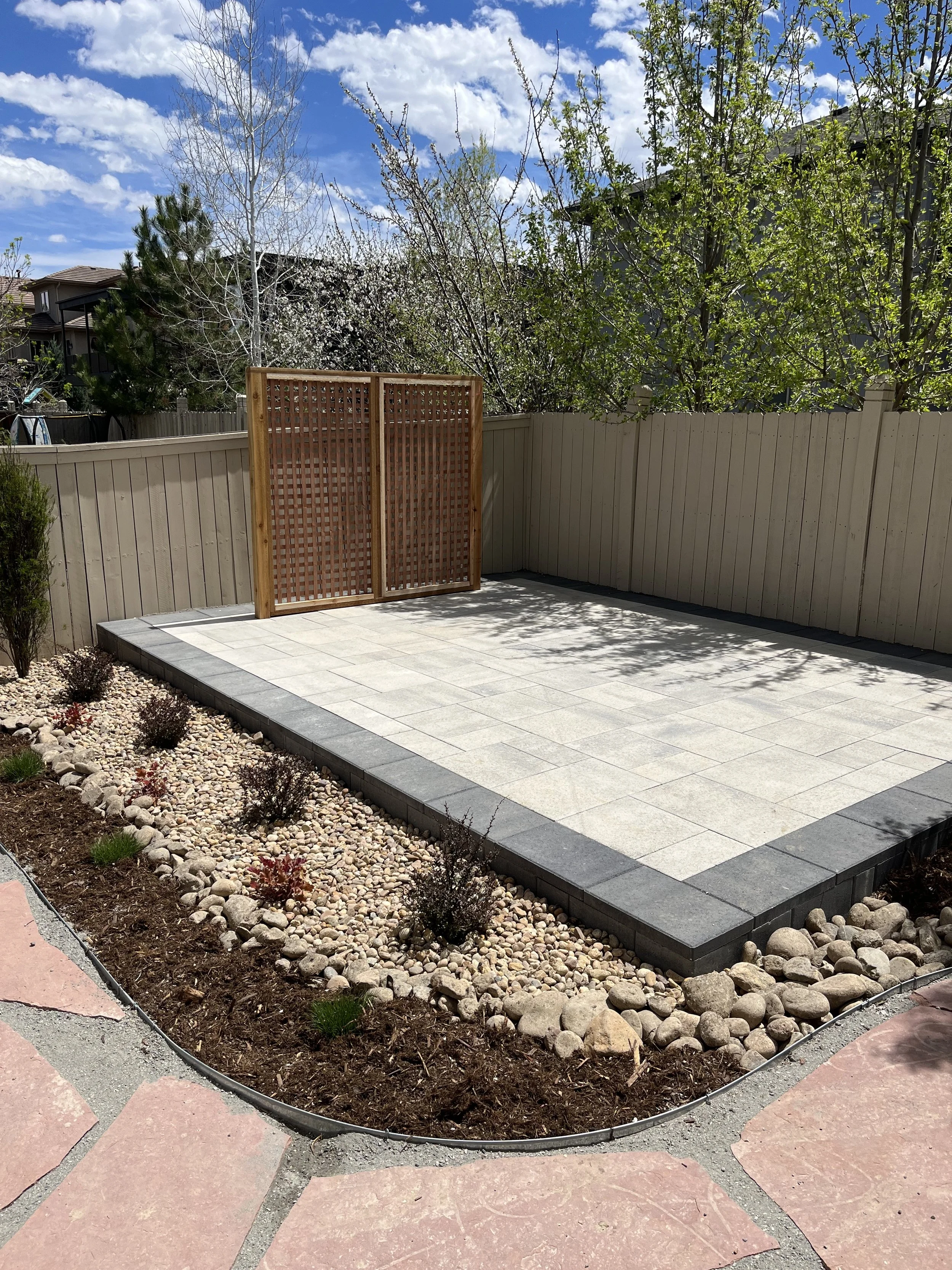 A backyard with a newly paved patio area, a wooden privacy screen, the flower bed with rocks and small plants, and trees and bushes in the background under blue skies with scattered clouds.