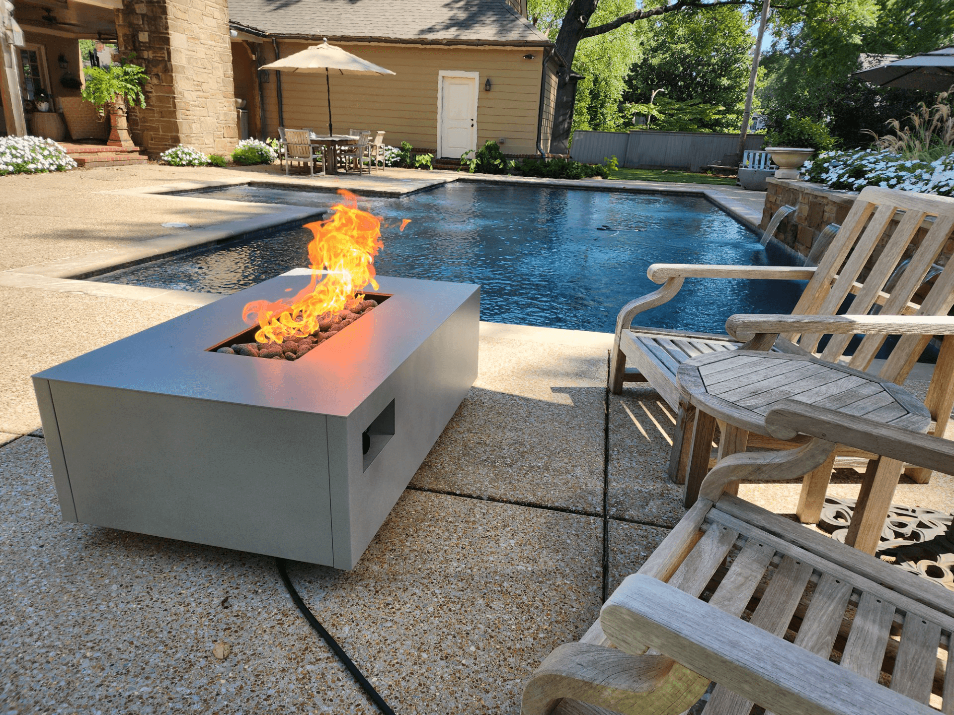 A backyard with a swimming pool, wooden chairs, a rectangular fire pit, a table with an umbrella, lush green trees, and flowering plants.