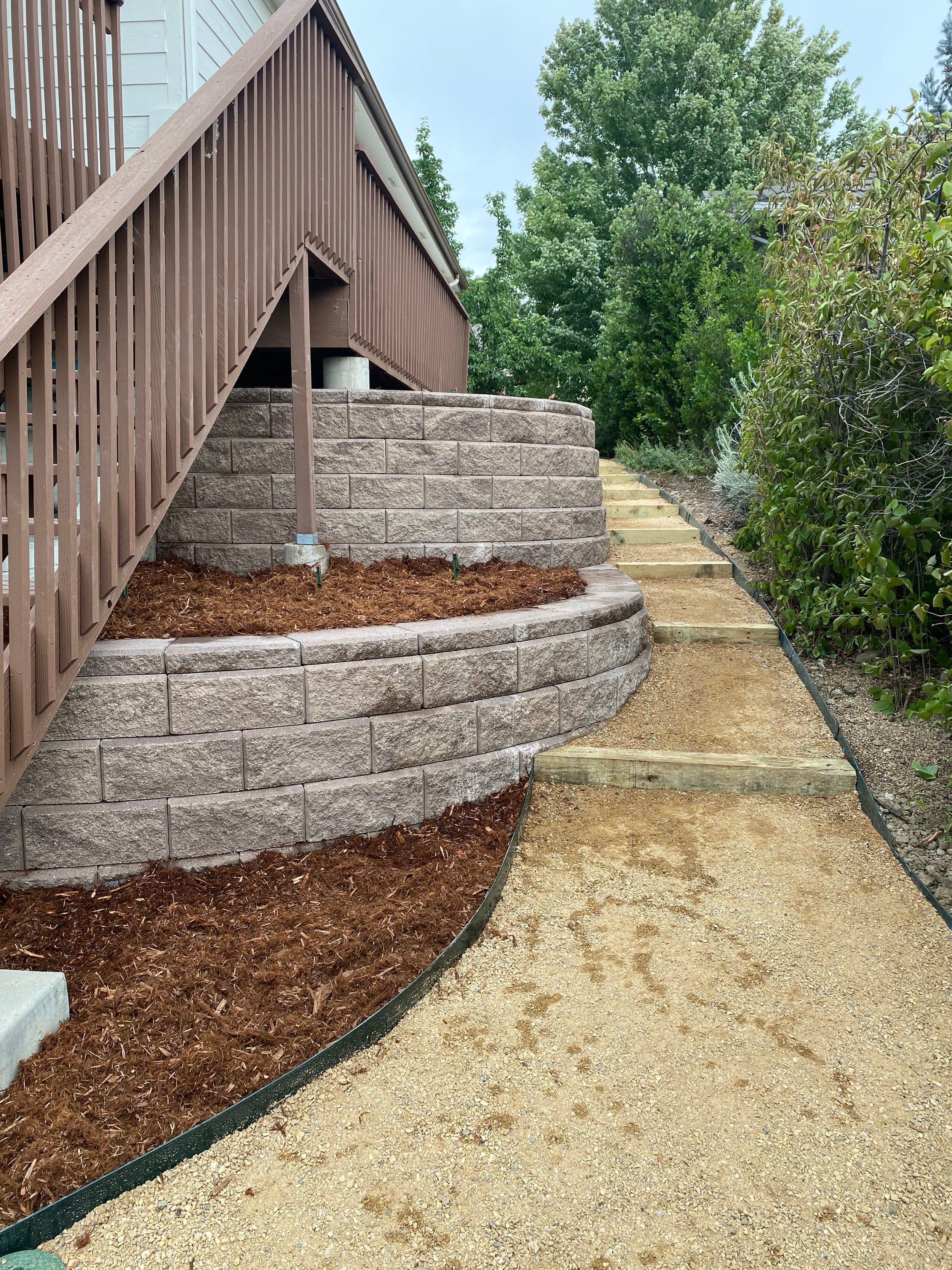 A garden pathway bordered by a retaining wall made of stone blocks, with a brown wooden deck and stairs on the left, mulch in a garden bed on the left, and a dirt pathway with wooden steps on the right, surrounded by green trees and bushes.