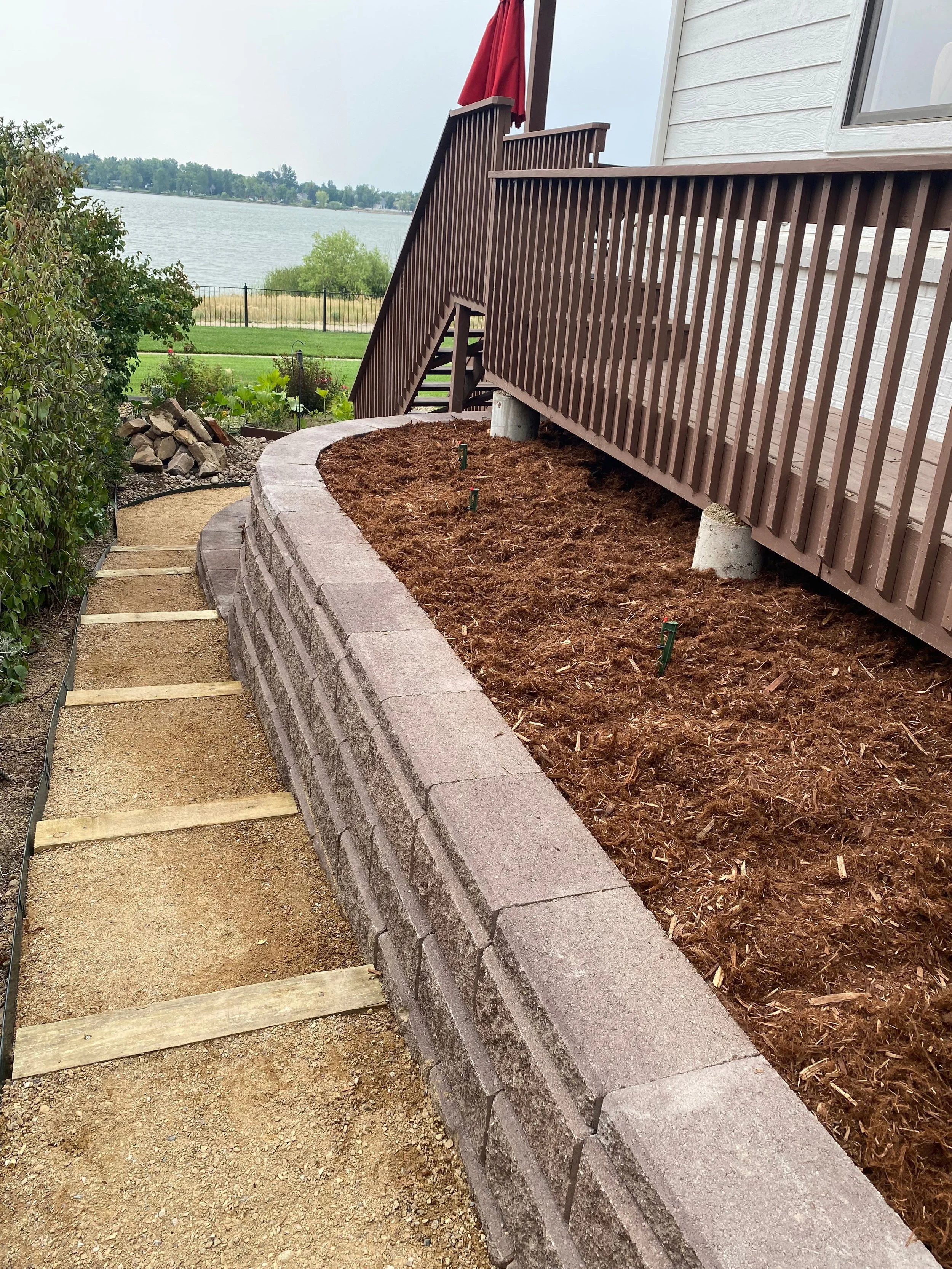 This image shows a landscaped backyard with a newly constructed retaining wall made of concrete blocks. There is a layer of soil behind the wall, likely prepared for planting. Alongside, a wooden deck with a staircase is attached to the house, overlo