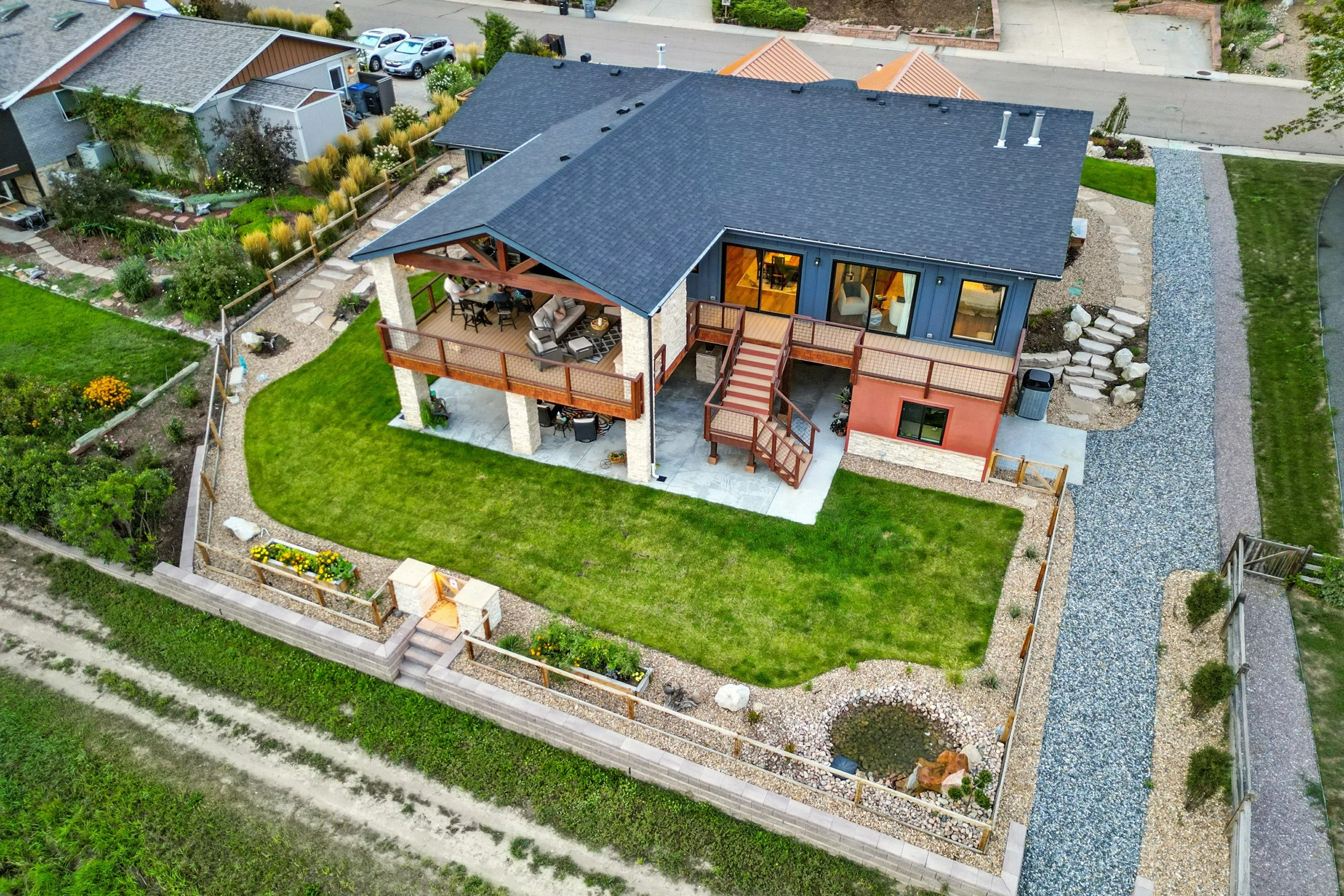 Aerial view of a modern two-story house with a dark blue roof, a large backyard with a lawn, outdoor patio with furniture, and surrounding landscaping, including a gravel driveway and flower beds.