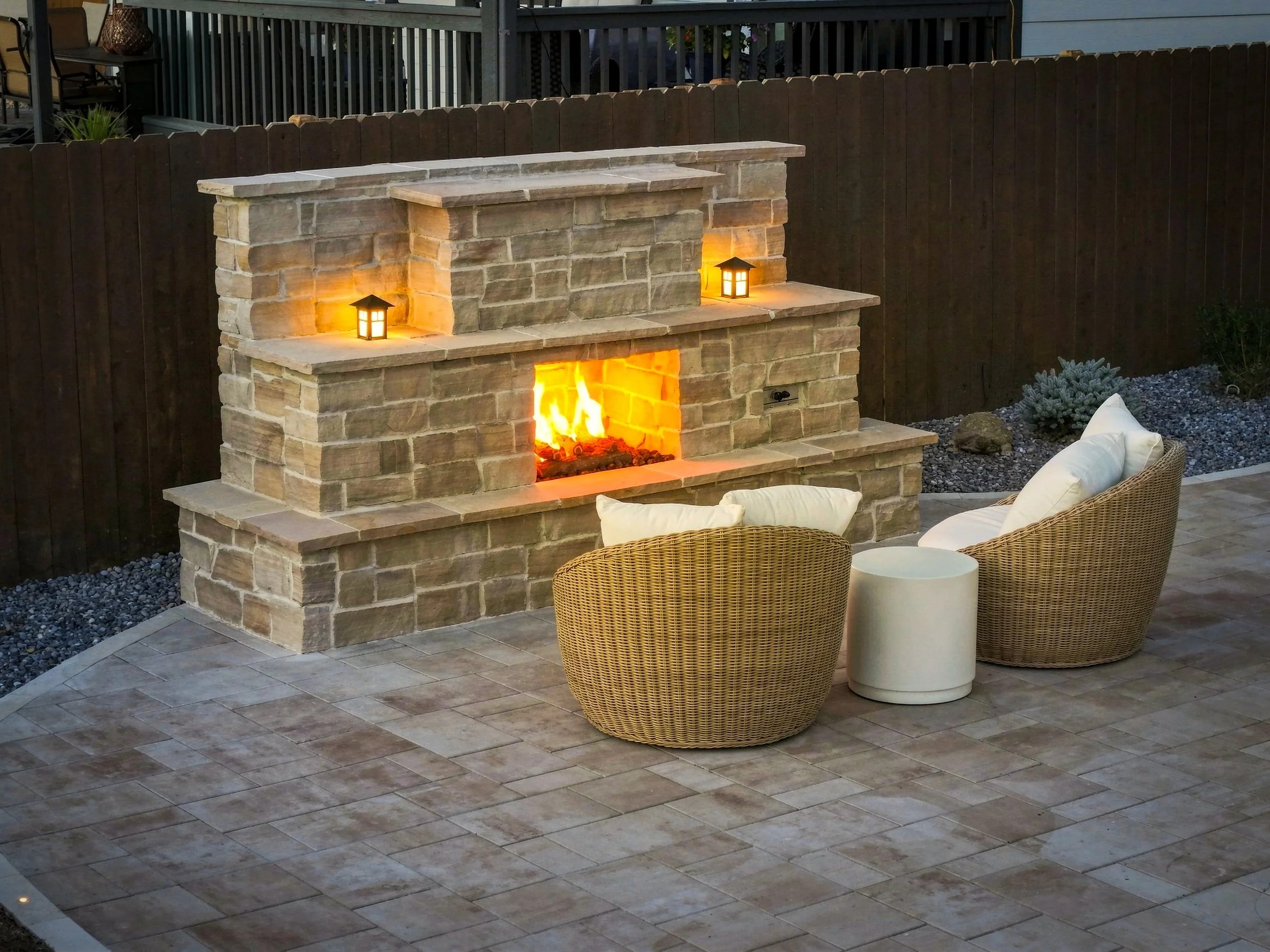 An outdoor patio area featuring a stone fireplace with two small lanterns on the mantel, surrounded by wicker chairs with cushions, a small white round table, and a wooden fence in the background.