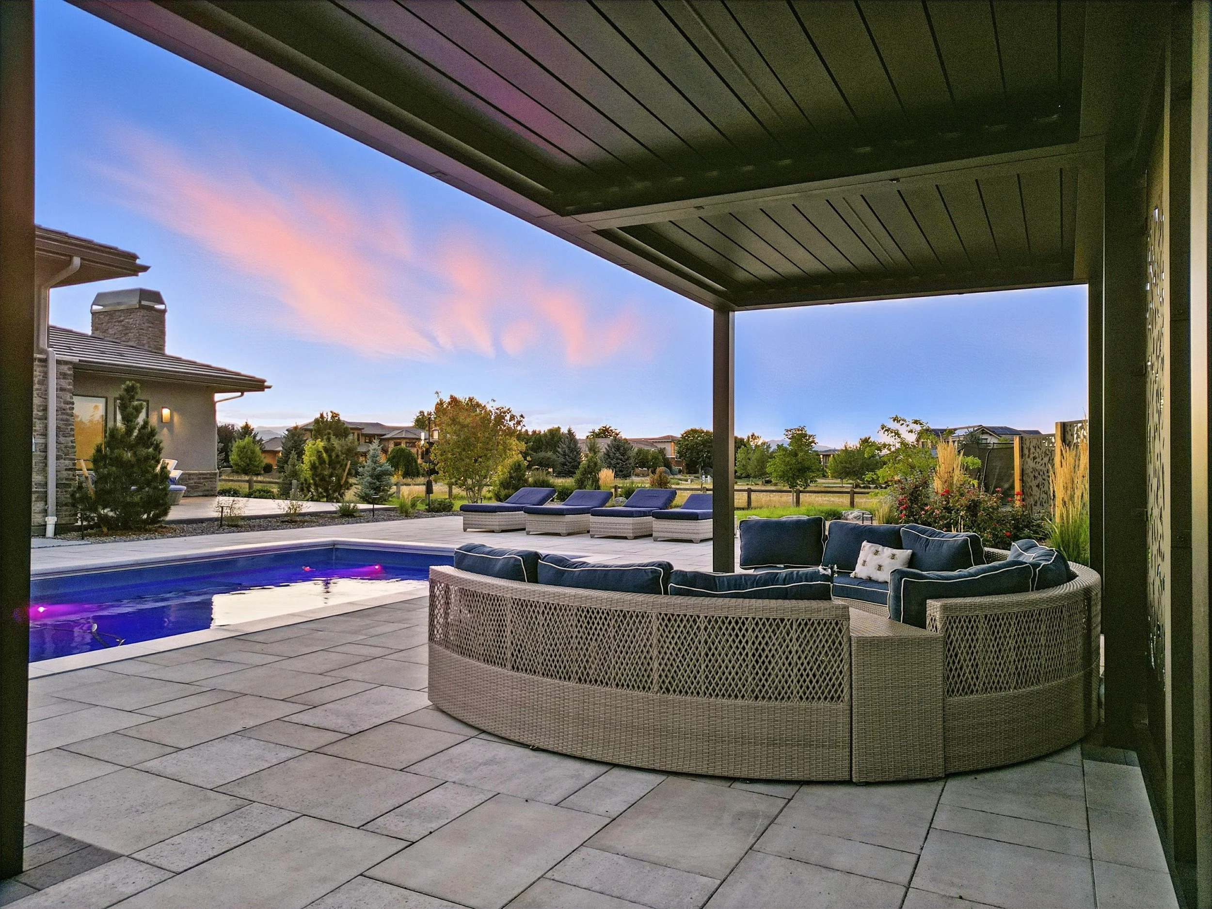 View of a backyard patio with a hot tub, sun loungers, and a sectional outdoor sofa, overlooking a grassy yard and trees at sunset.