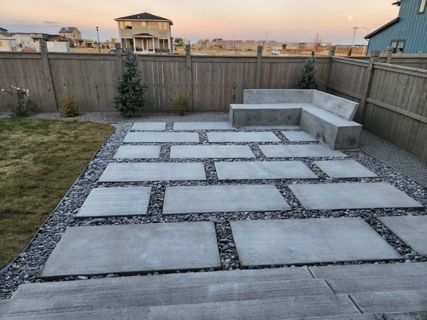 Backyard patio with large rectangular concrete pavers, smaller round river rocks, and a built-in concrete bench, enclosed by a wooden fence with small trees and shrubs outside.
