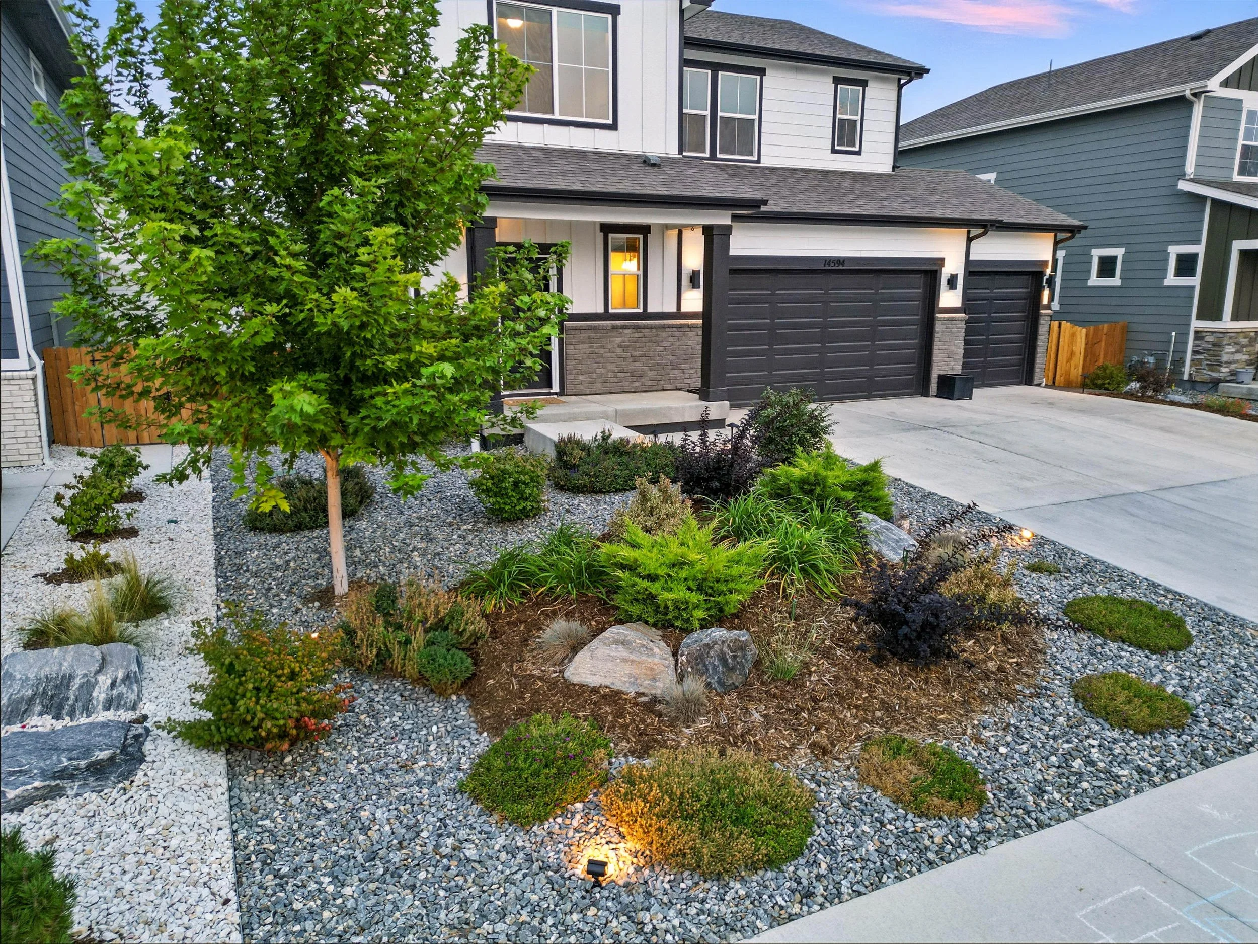 A modern two-story house with a gray and black exterior, a driveway, and a landscaped front yard with various green plants, a small tree, and rocks.