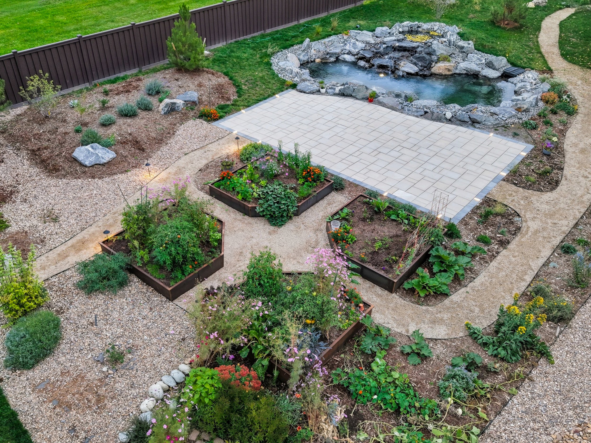 A landscaped backyard garden with a pond surrounded by rocks, a tiled patio area, pathways, and various flower beds with plants and flowers.