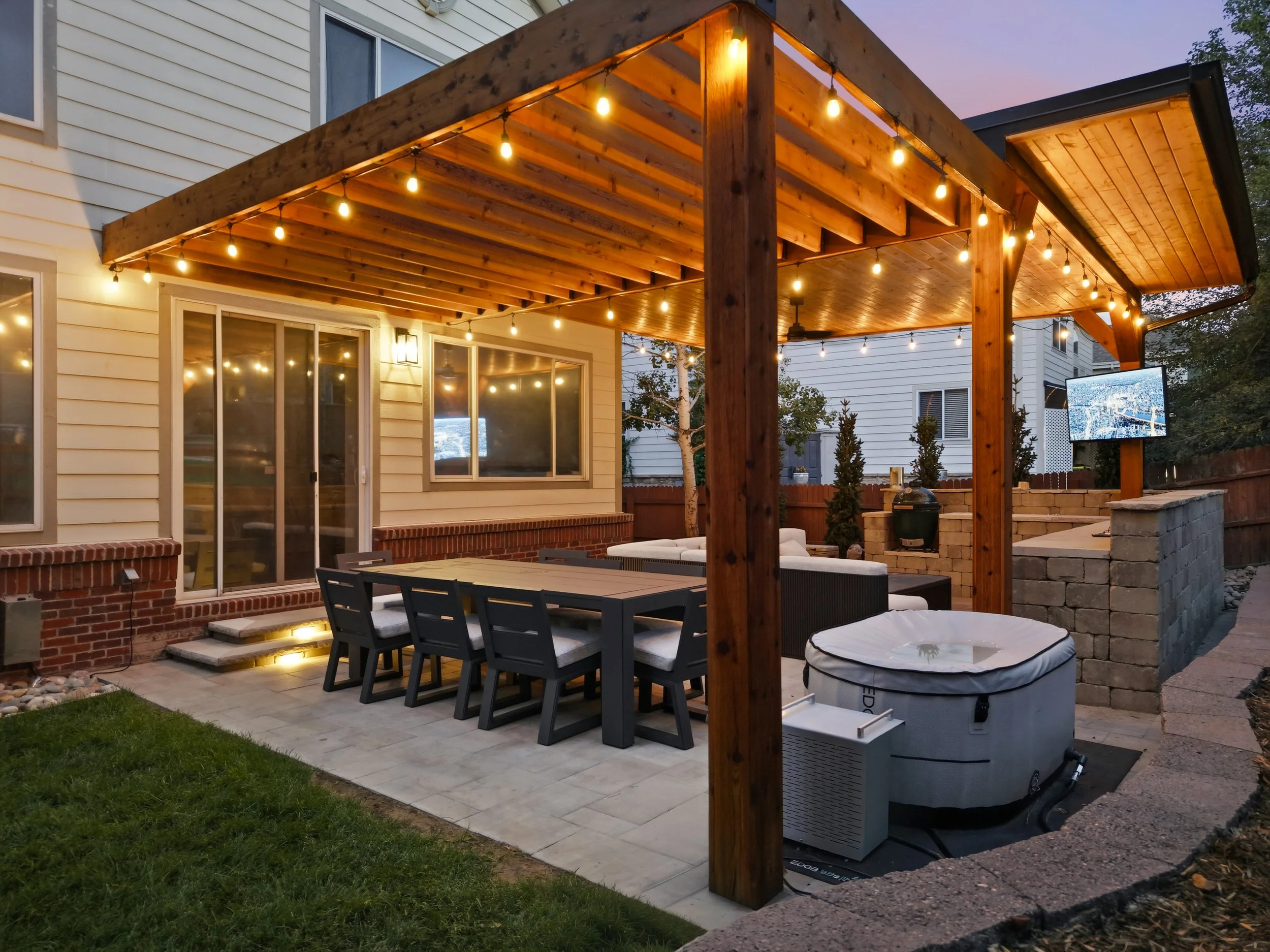 Backyard patio with a wooden pergola decorated with string lights, a dining table with chairs, a television on the side, and a brick and stone BBQ grill, during dusk.