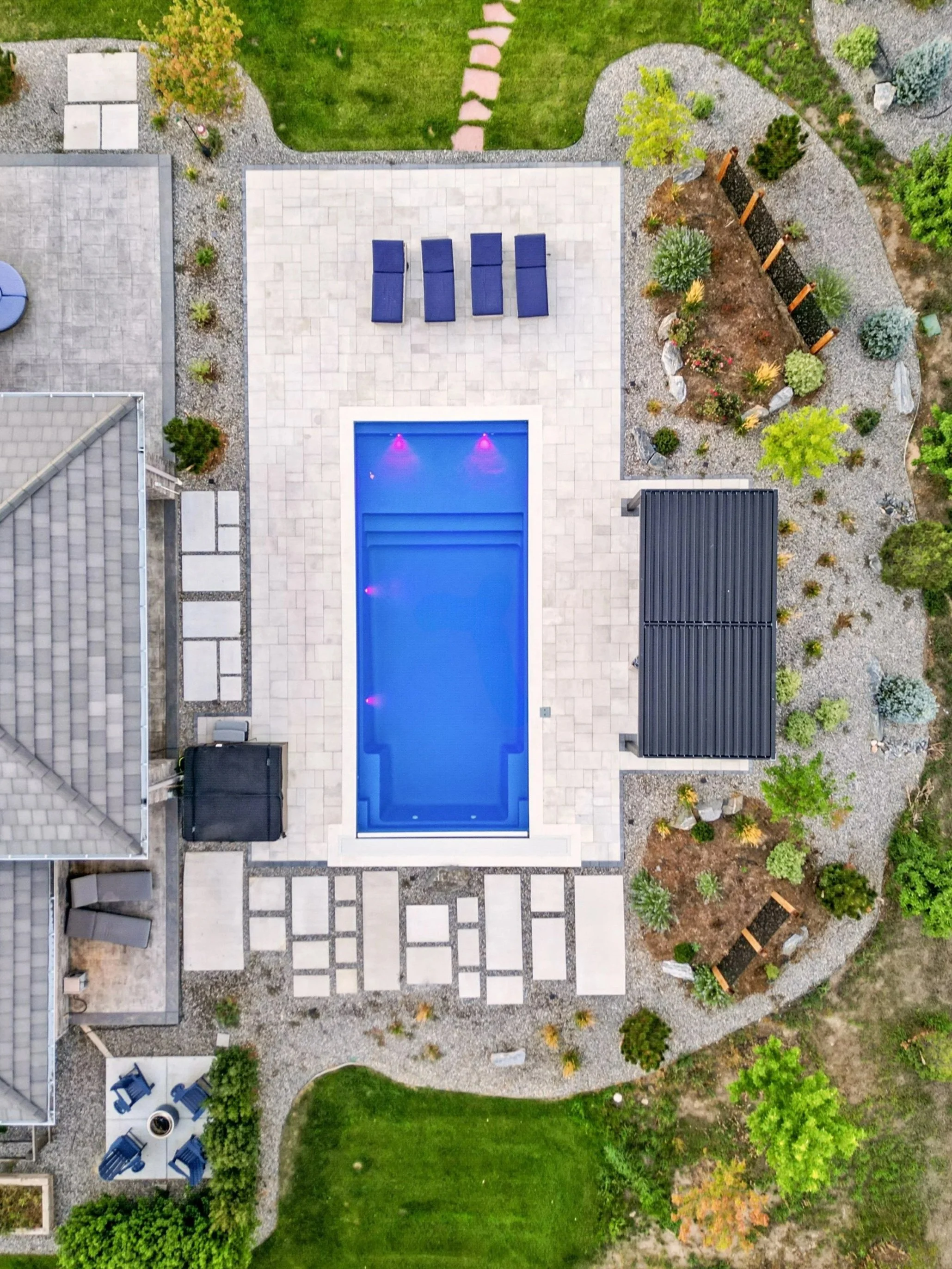 A backyard swimming pool, surrounded by a patio with lounge chairs, a garden area with plants and trees, and a house with a gray roof.