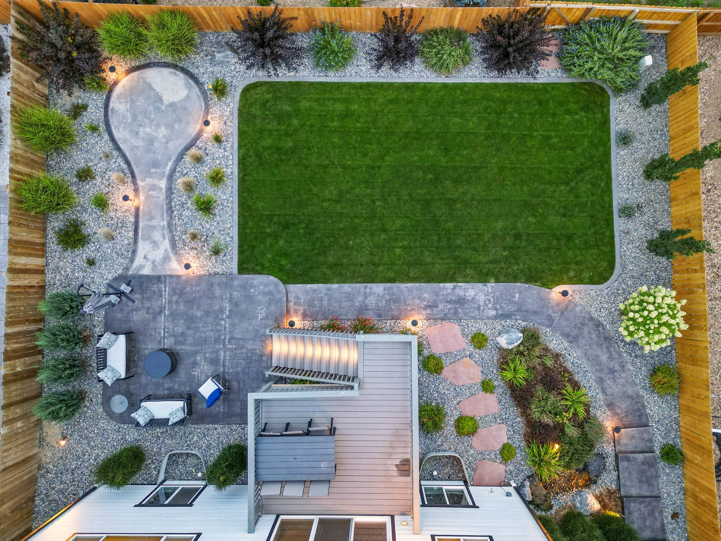 Aerial view of a landscaped backyard featuring a rectangular green lawn, a curved stone pathway with outdoor seating, small trees and plants, and a wooden fence surrounding the yard.