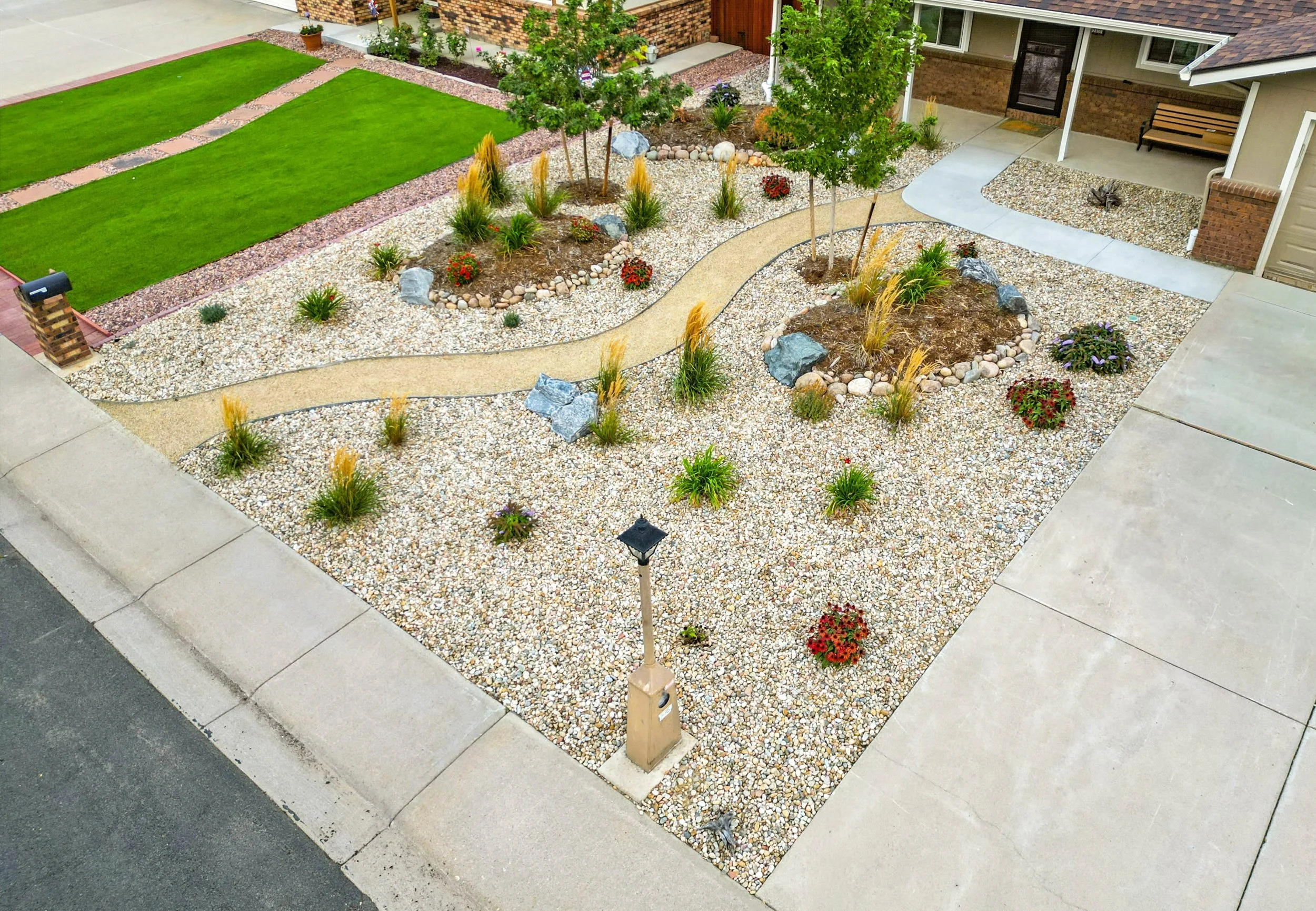 View of a landscaped front yard with a curved walkway, green lawn, small trees, mulch beds with rocks and plants, a lamp post, and a house with a porch and garage.