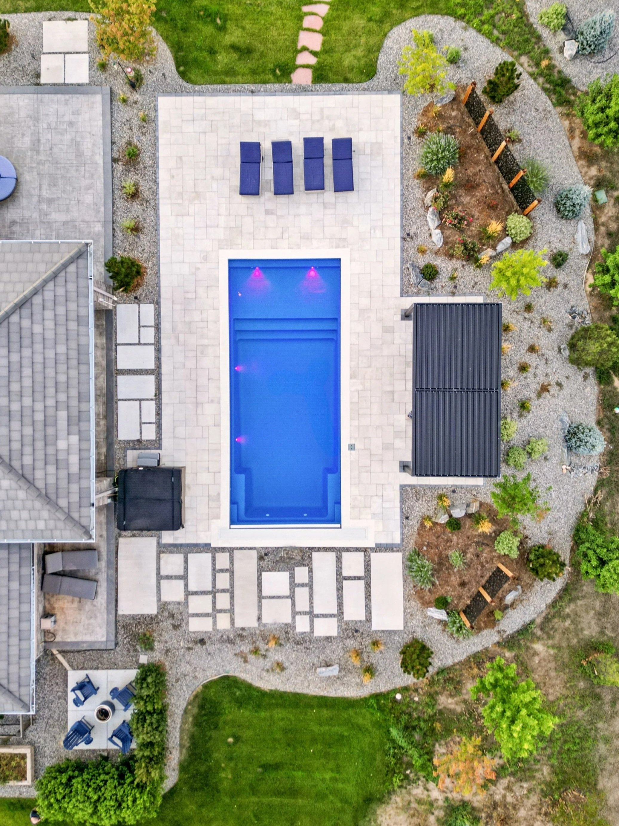 Aerial view of a backyard with a swimming pool, surrounded by a paved patio, lounge chairs, garden beds, and lush green grass.