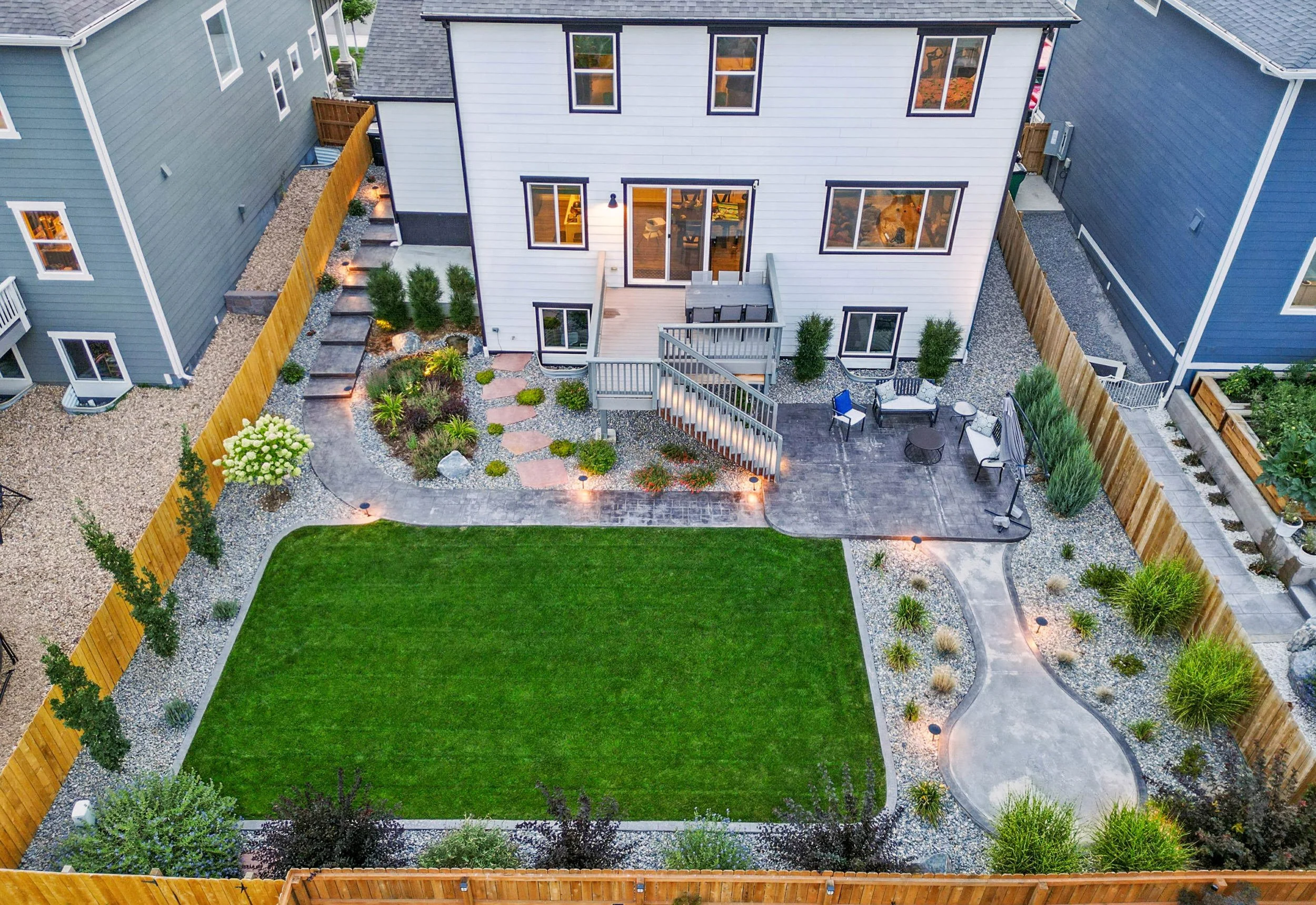 A backyard view showing a green lawn, a paved patio area with outdoor seating, a landscaped garden with plants and trees, and a two-story house with large windows. The yard is enclosed by a wooden fence and features pathway lighting.