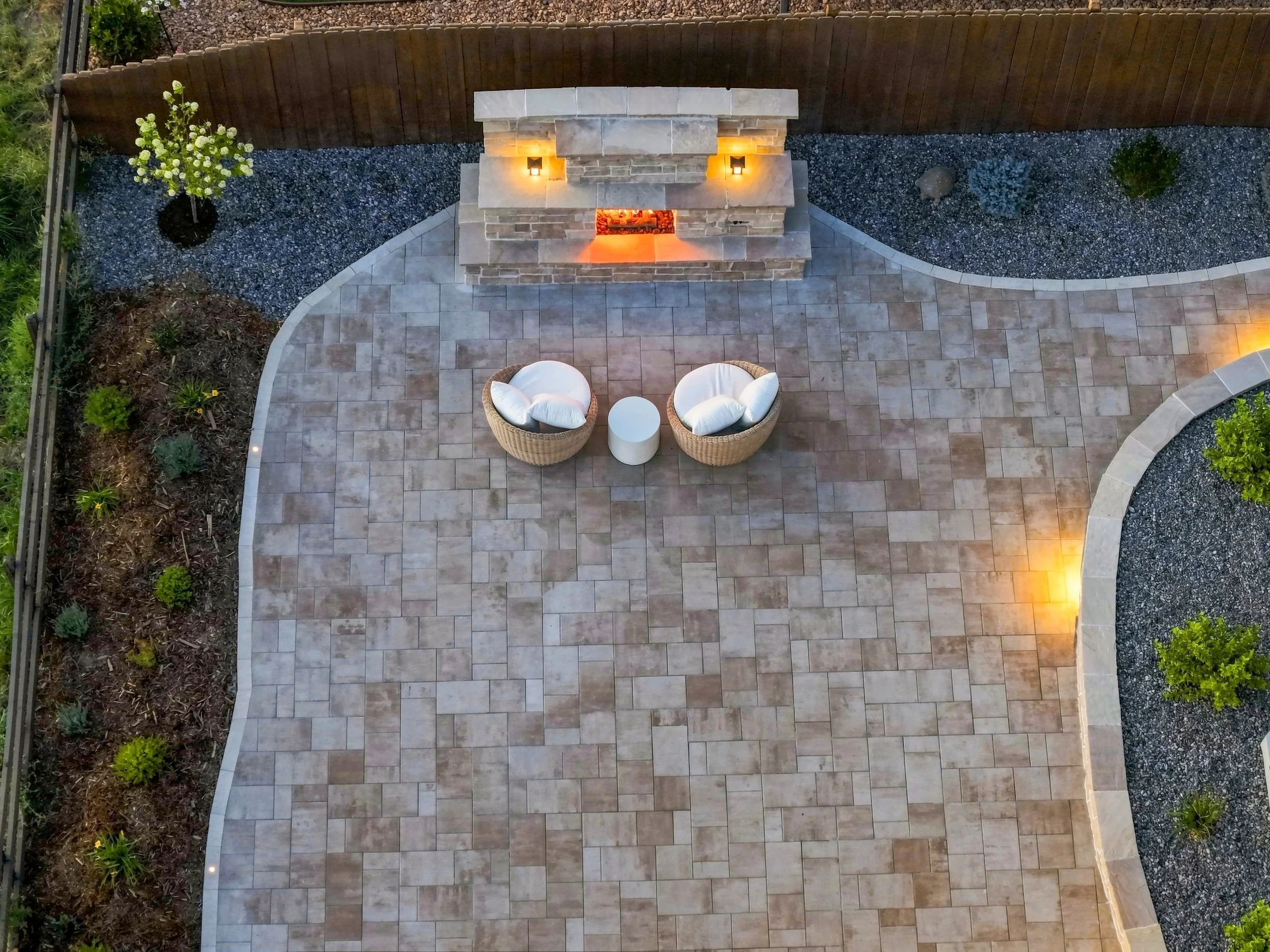A backyard patio with a beige tiled floor, a stone fireplace with two wall-mounted lights and a fire burning inside, and wicker chairs with white cushions.