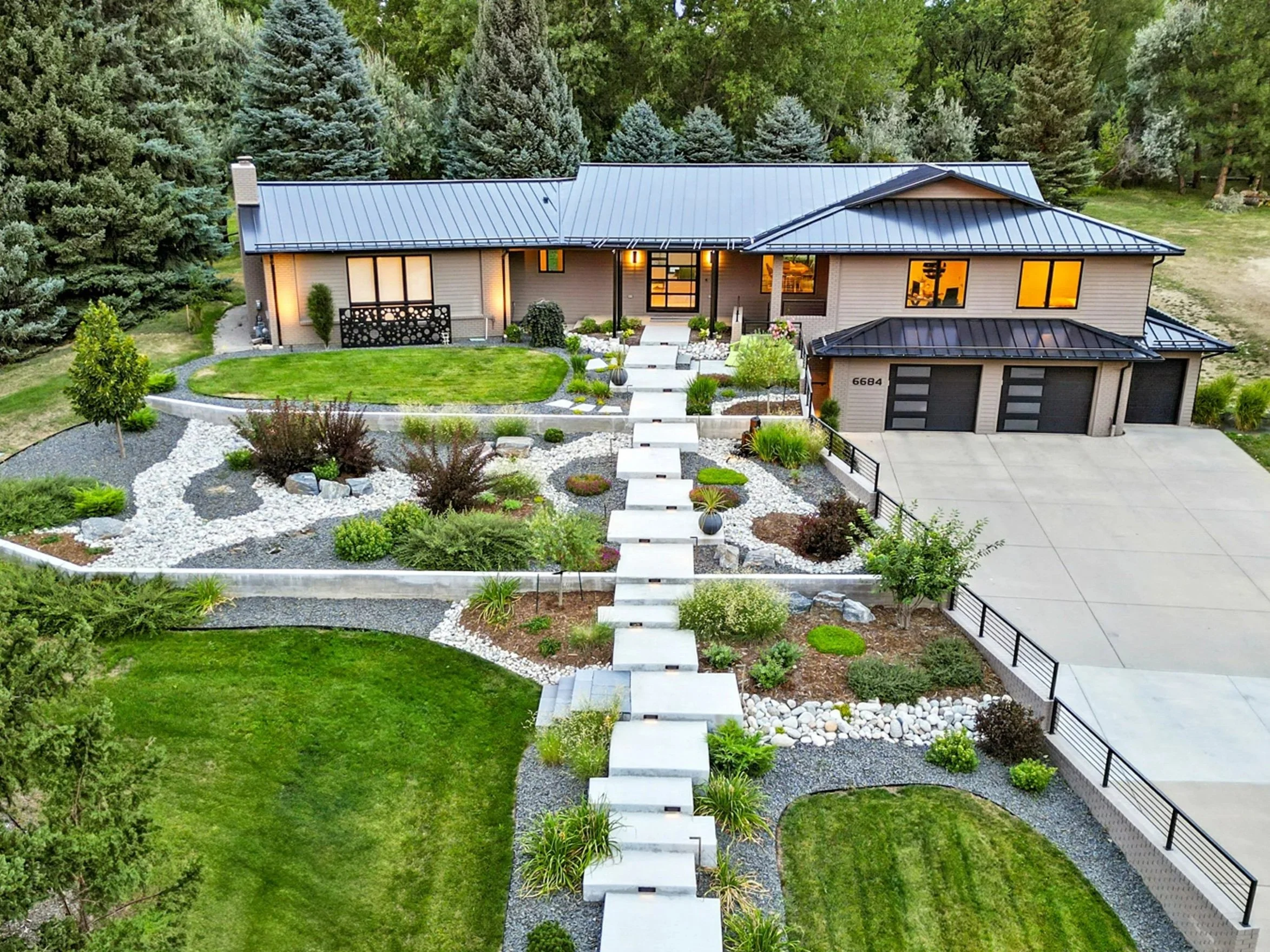 A modern house with a metal roof, surrounded by well-maintained lawn and landscaped garden with rocks and small trees, with a large concrete driveway and a pathway leading to the front door.