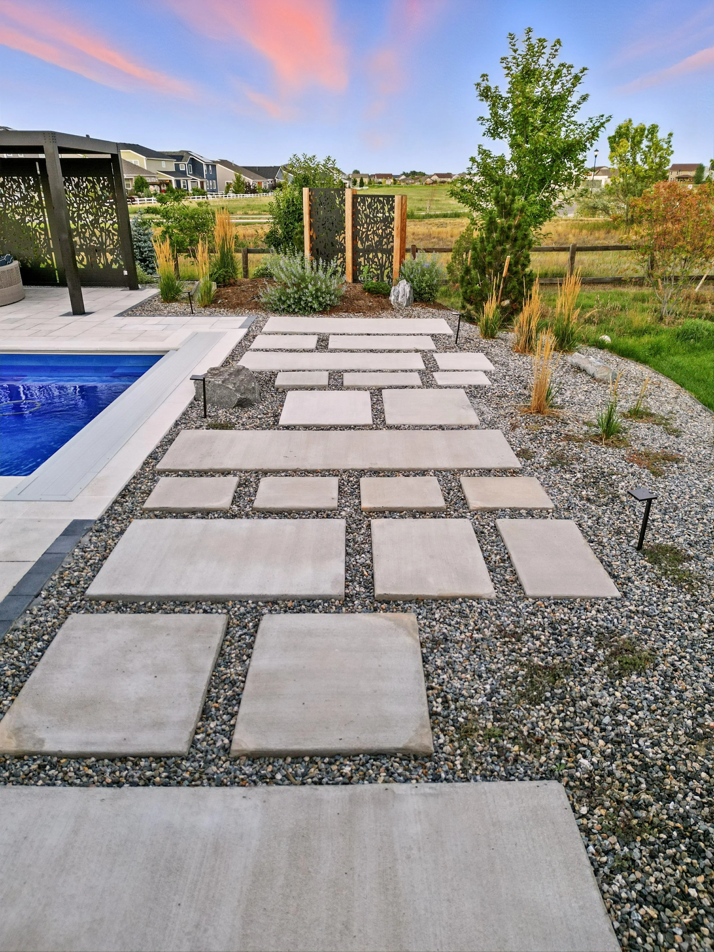 A backyard with a swimming pool on the left, a stone pathway, landscaped plants, a wooden fence, and a view of open grassy fields and houses in the background under a colorful sunset sky.