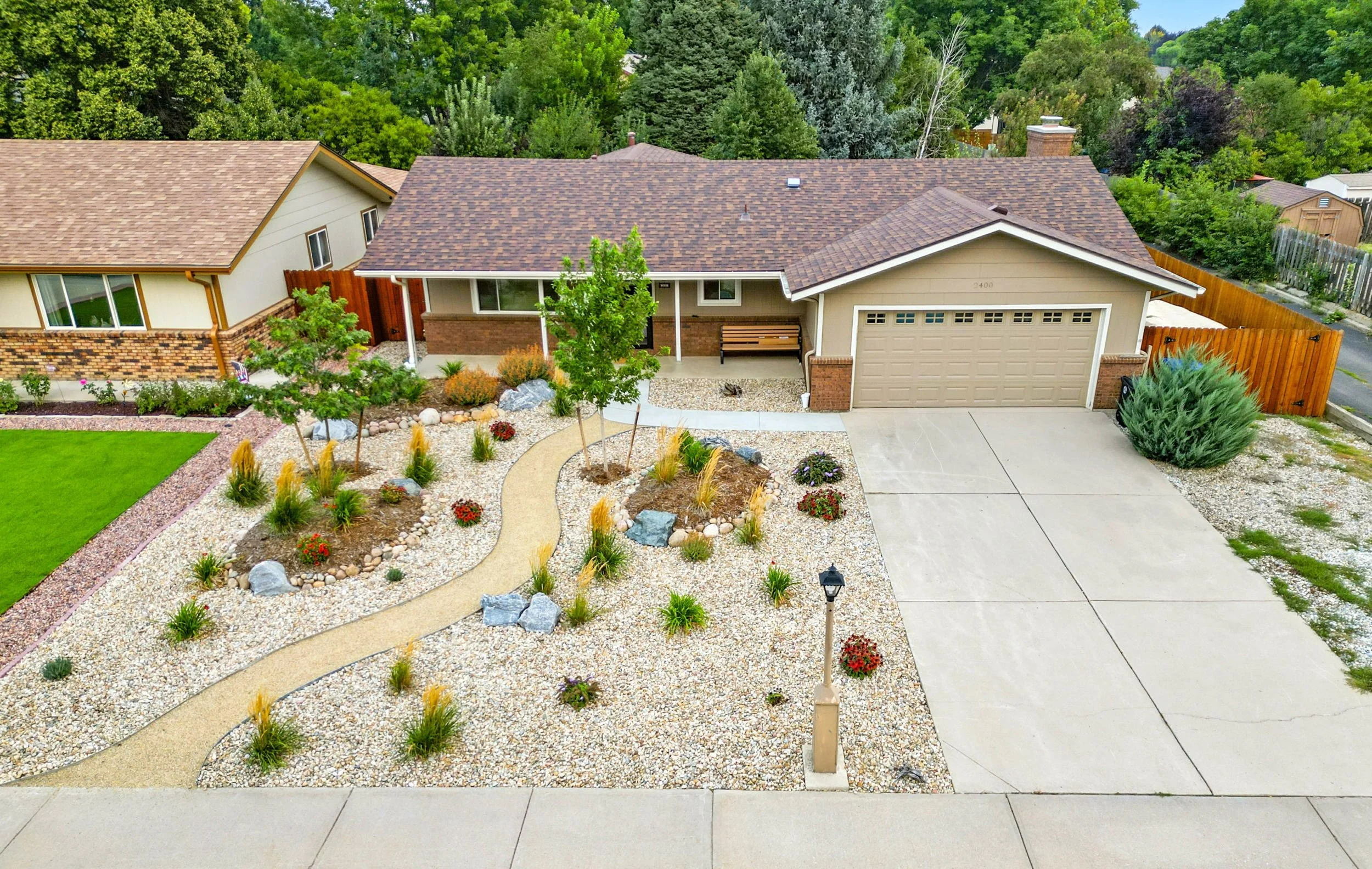 Aerial view of a suburban house with a landscaped front yard featuring a gravel garden, small trees, flowering plants, a curved pathway, and a concrete driveway.