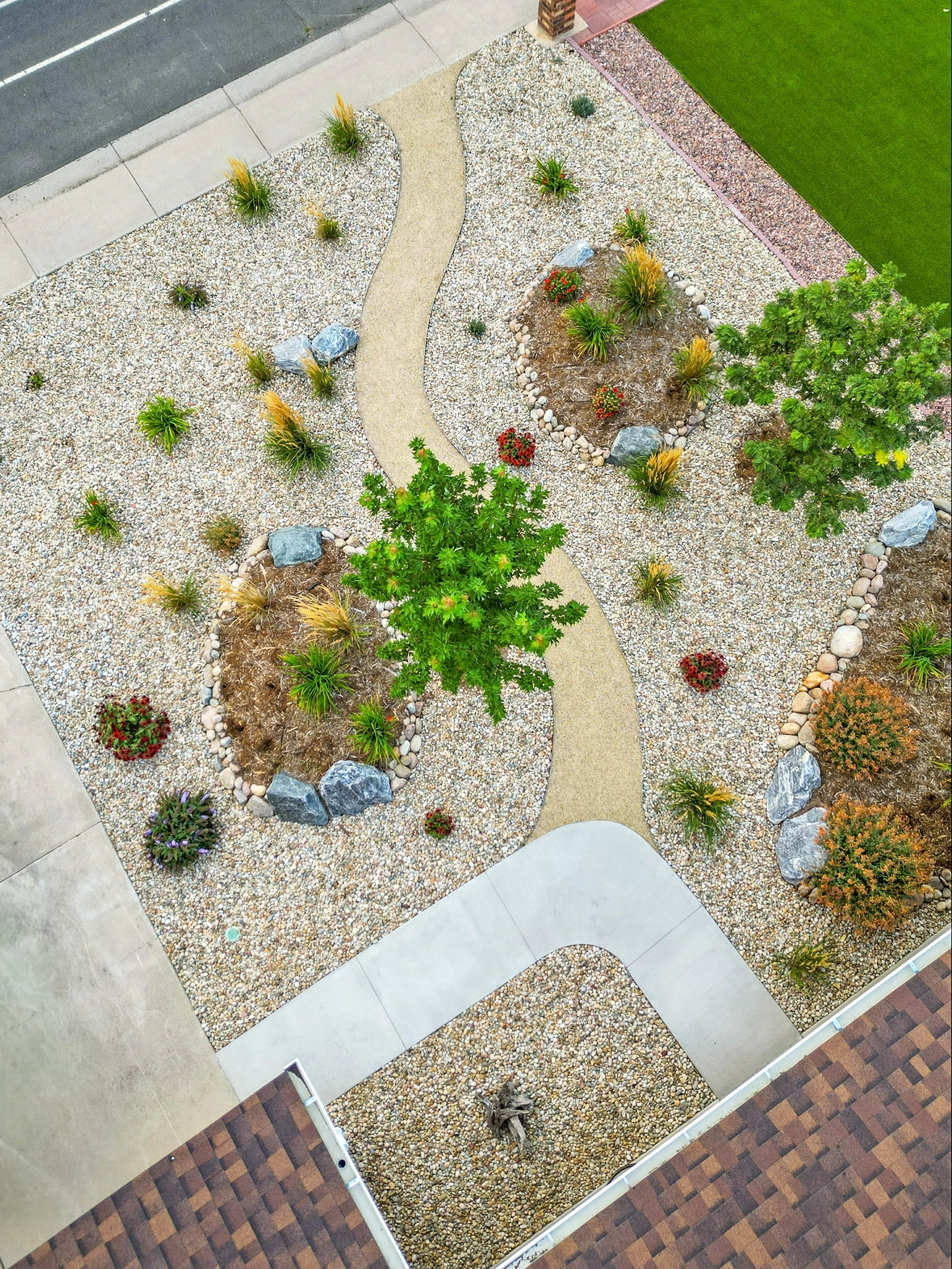 An aerial view of a landscaped yard with a winding concrete pathway, small bushes, ornamental grasses, rocks, decorative mulch, flowering plants, and surrounding sidewalk and brick patio areas.