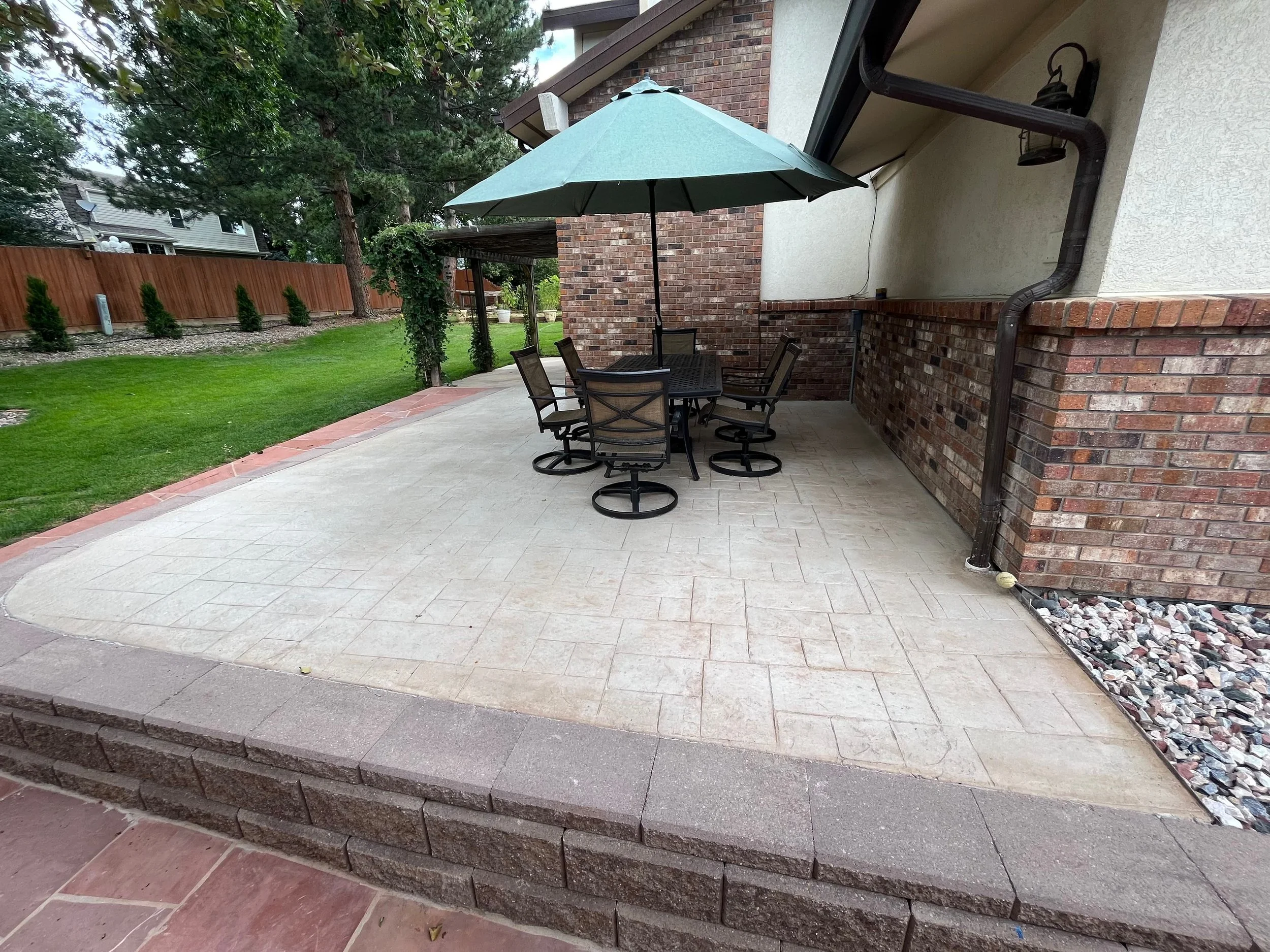 A backyard patio with a round table, six chairs, and a large green umbrella. The patio has a brick and concrete surface, bordered by bricks, with a grassy yard, trees, and a wooden fence in the background.