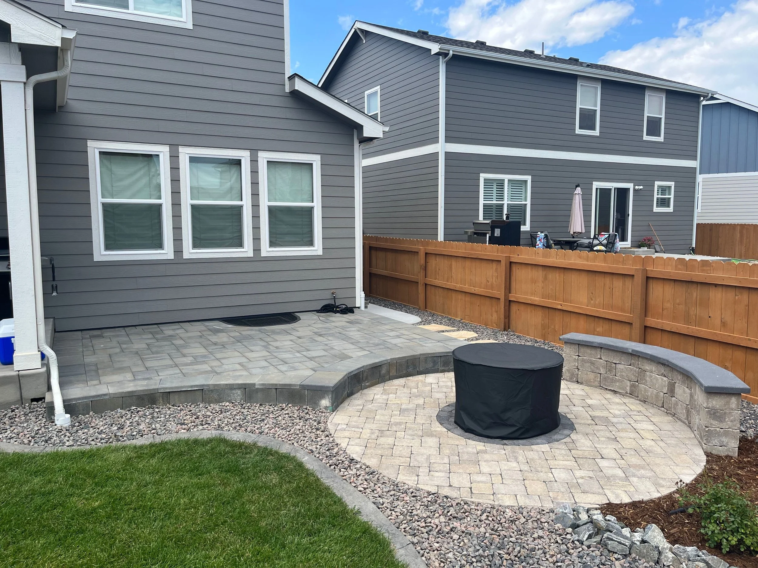 Backyard patio area with gray pavers, a curved stone wall, a covered round table, and a wooden privacy fence, adjacent to a gray house with multiple windows.