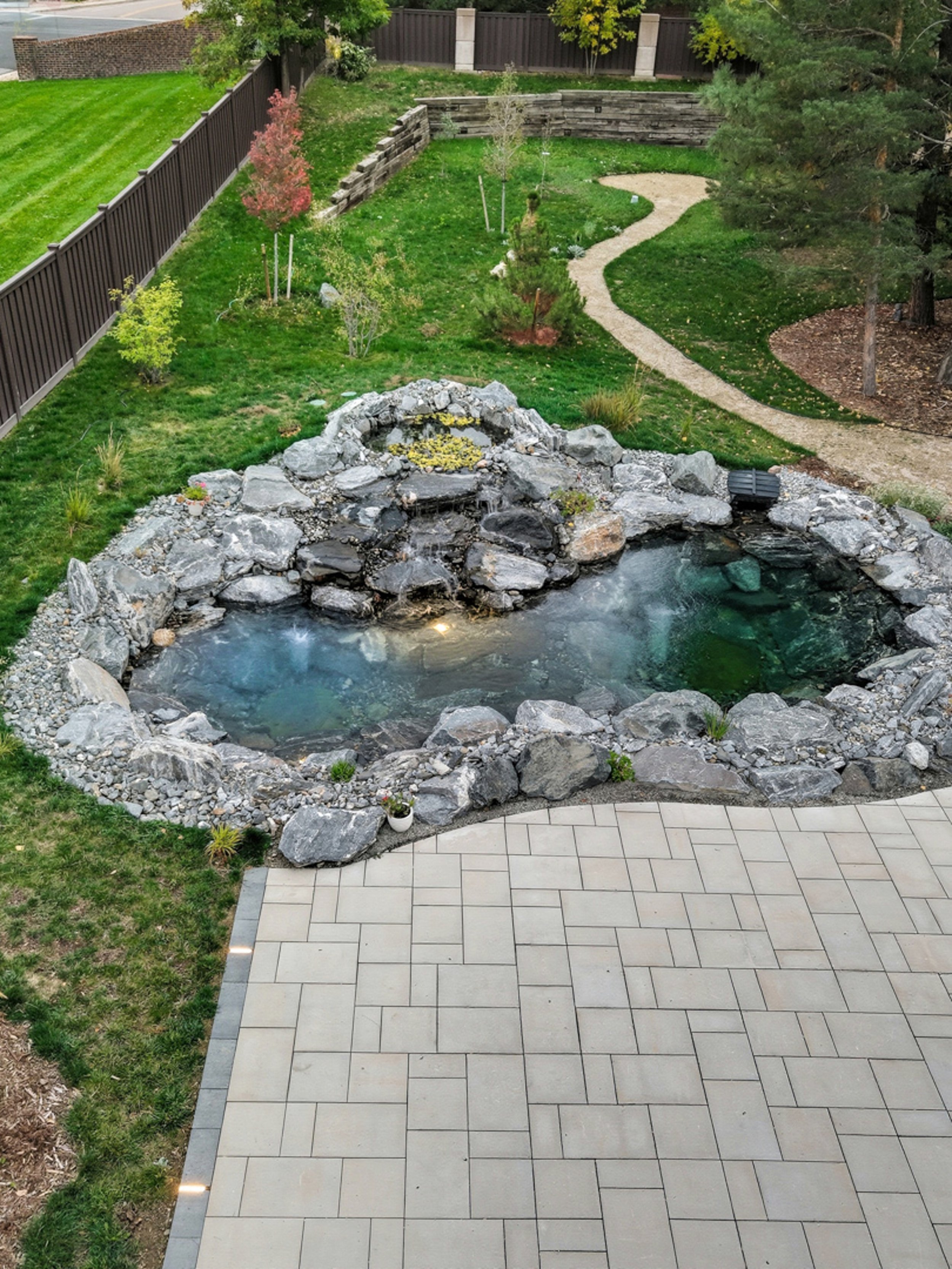 A backyard with a stone-lined hot tub with water, surrounded by a patio, lawn, and a curved walkway, with trees and a wooden fence in the background.