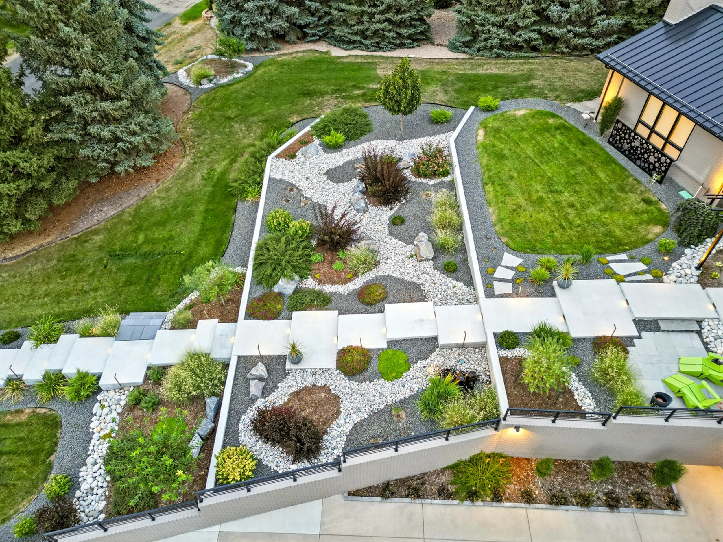 Aerial view of a modern backyard garden with a small lawn, decorative rock garden, lush trees, and outdoor seating area with green lounge chairs.