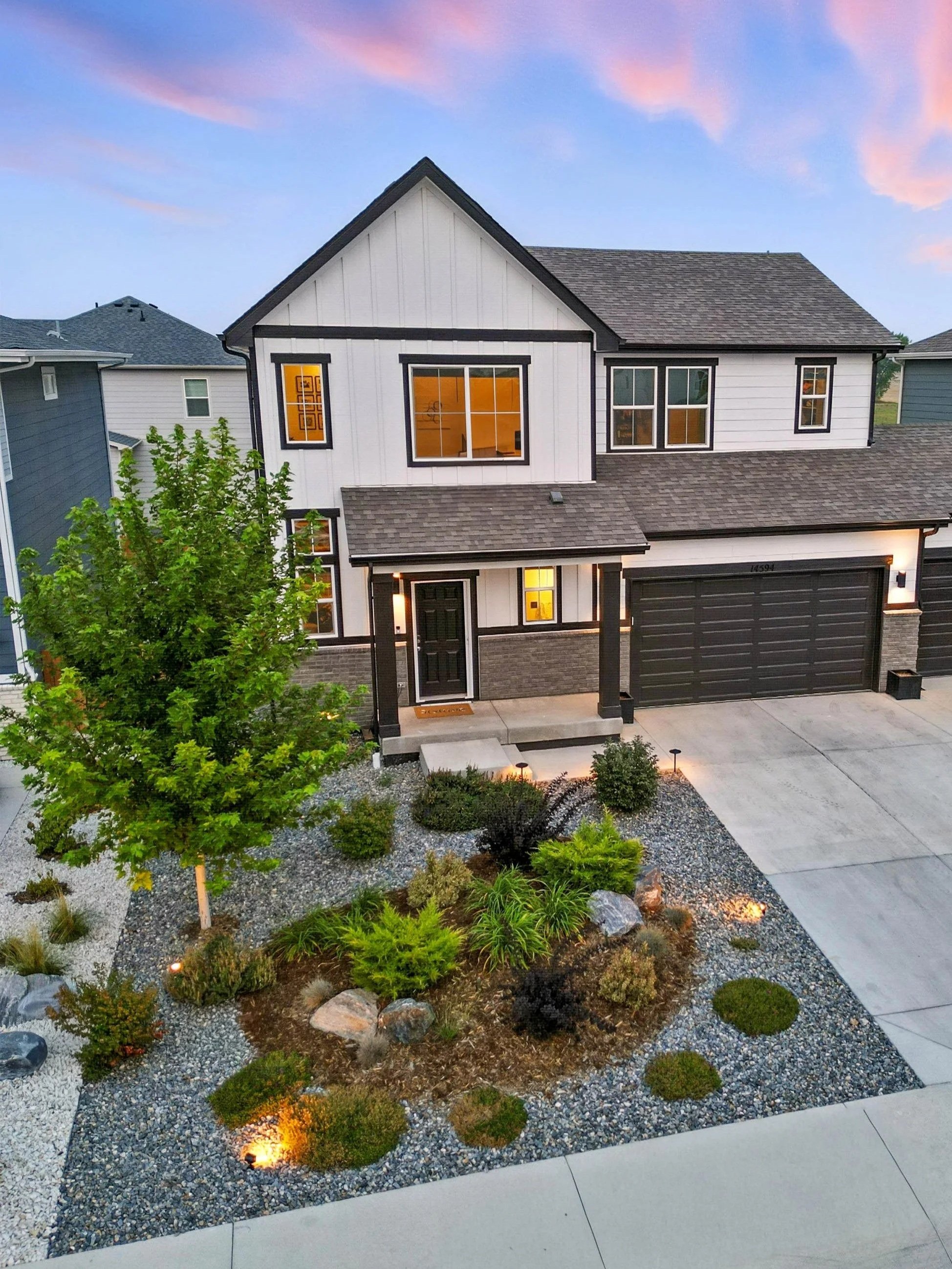 A modern two-story house with black trim, a front porch, a two-car garage, and a landscaped front yard with small bushes, a tree, and rocks, illuminated by outdoor lights at dusk.
