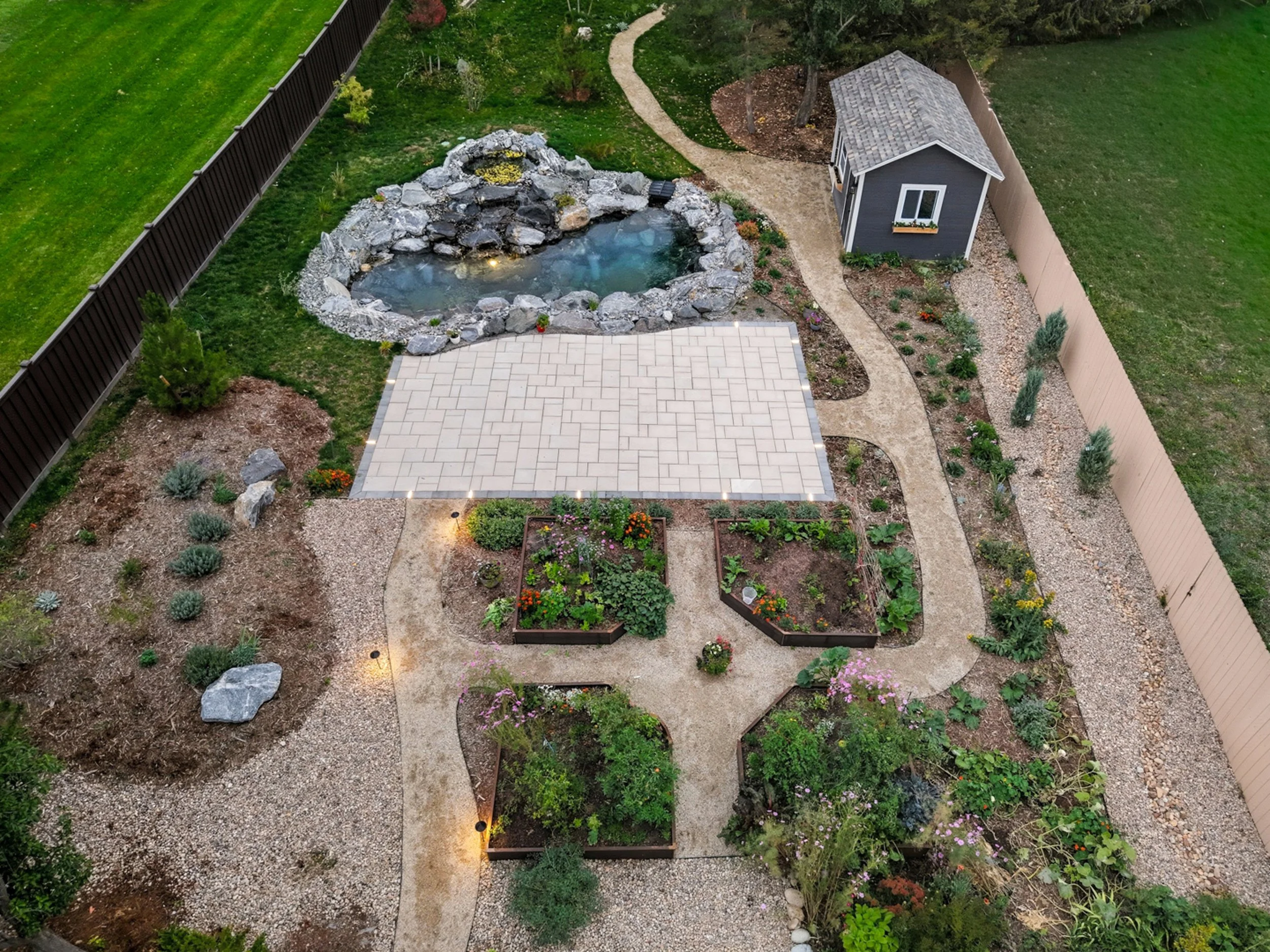 A backyard garden with a pond, a paved patio, vegetable beds, a small shed, and walking paths, surrounded by a wooden fence and lush green lawn.
