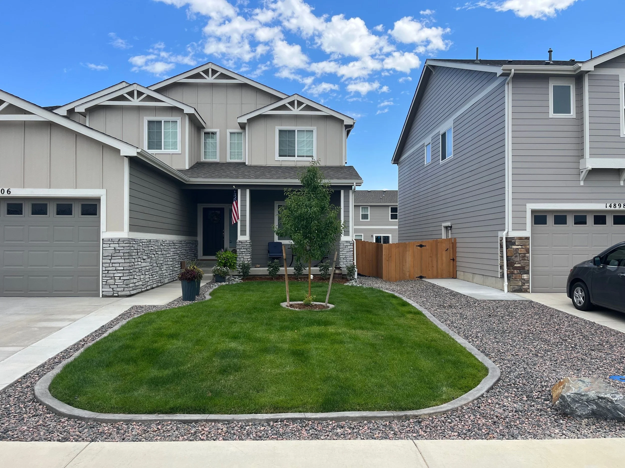 Front view of a modern two-story house with gray siding, stone accents, and a small front yard with grass and trees, flanked by neighboring houses. The driveway on the right has a parked car, and the sky is partly cloudy.