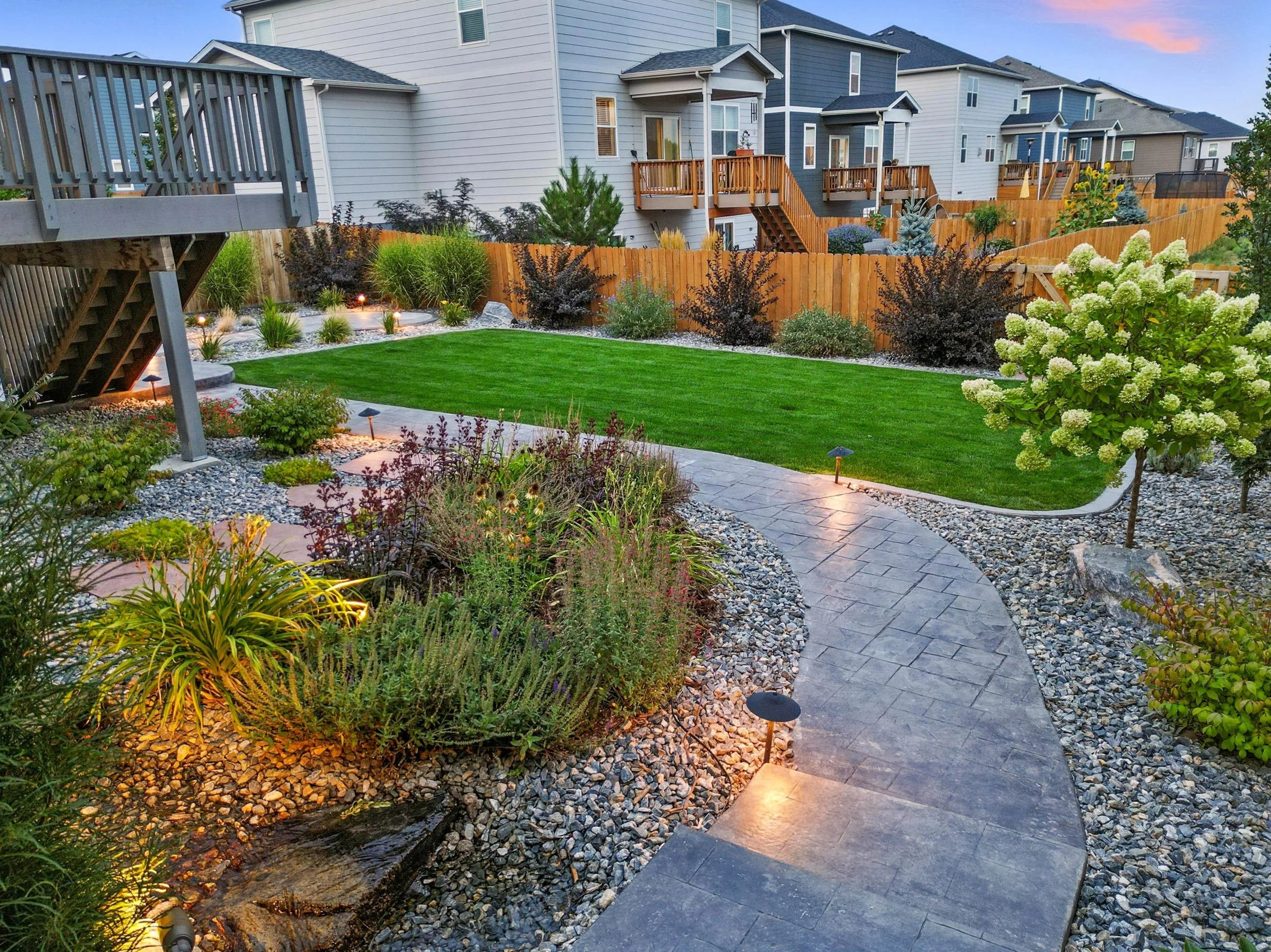 Backyard with manicured green lawn, curved stone pathway, decorative rocks, bushes, trees, and small outdoor lights, with neighboring houses in the background.