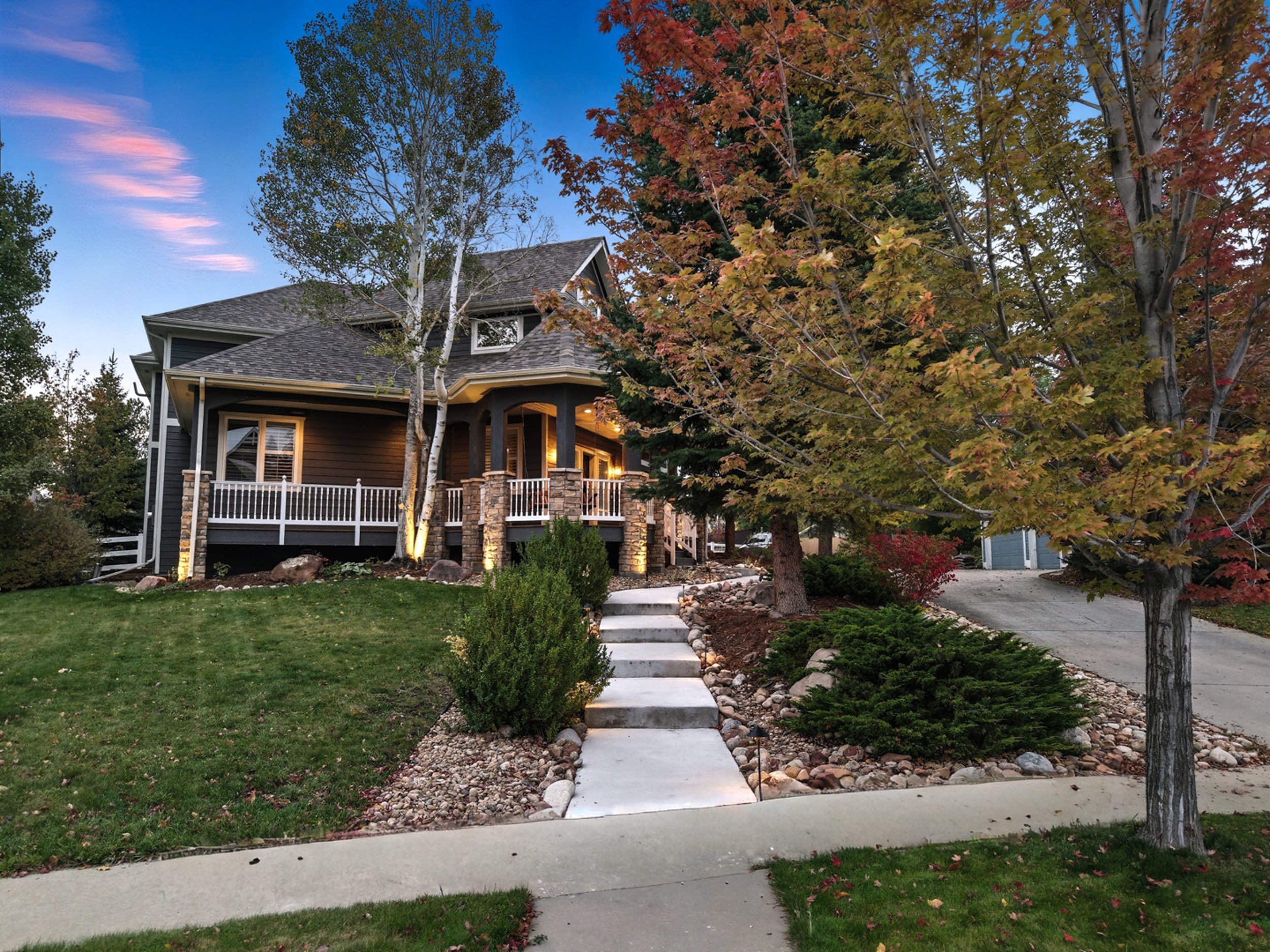 Front yard of a house with a sidewalk, landscaped with trees, bushes, and rocks, during twilight.