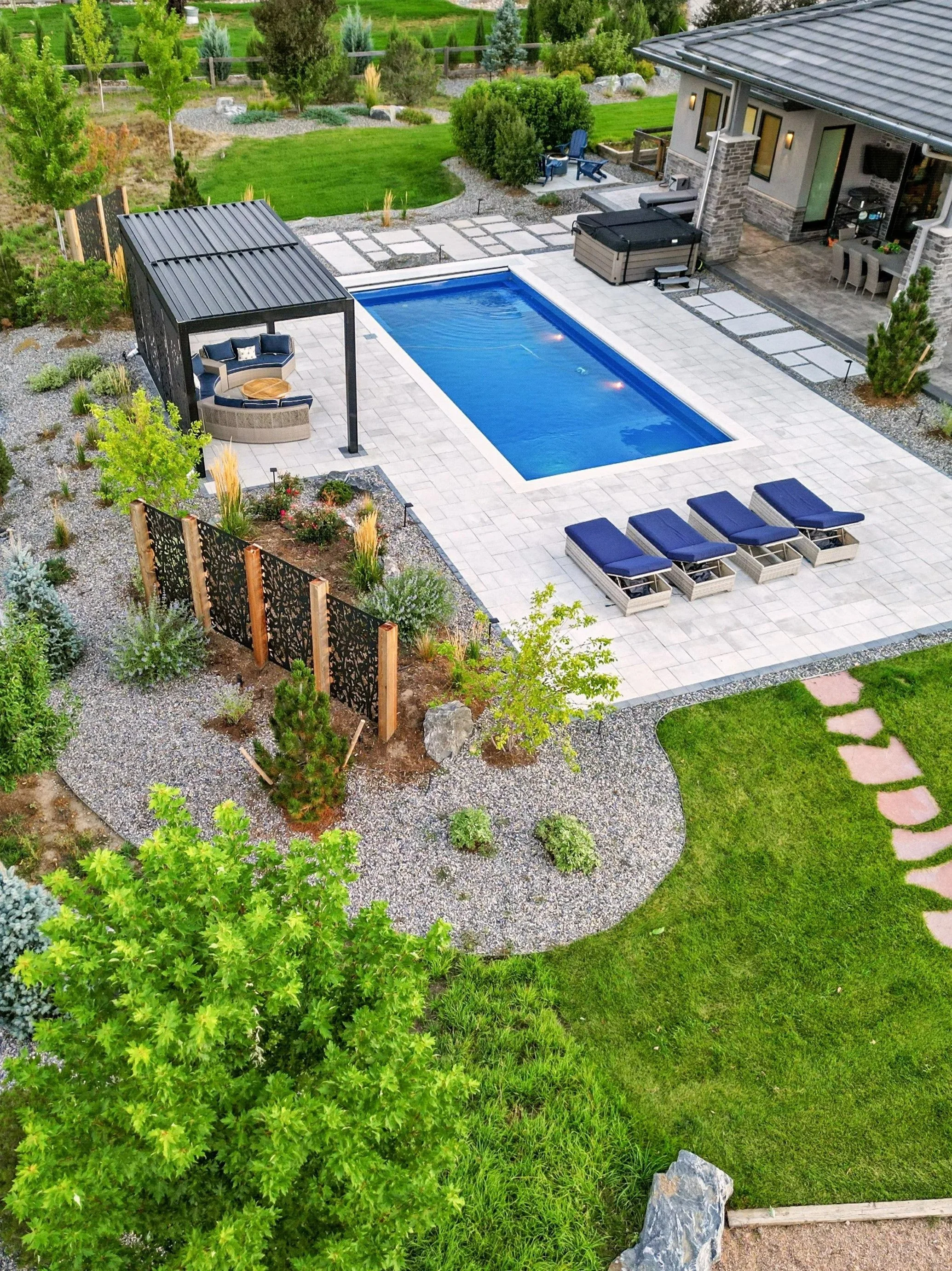 A backyard with a swimming pool, surrounded by a patio with lounge chairs, a gazebo, a hot tub, and outdoor furniture. The yard has green grass, trees, shrubs, and a garden area with plants and a pathway.