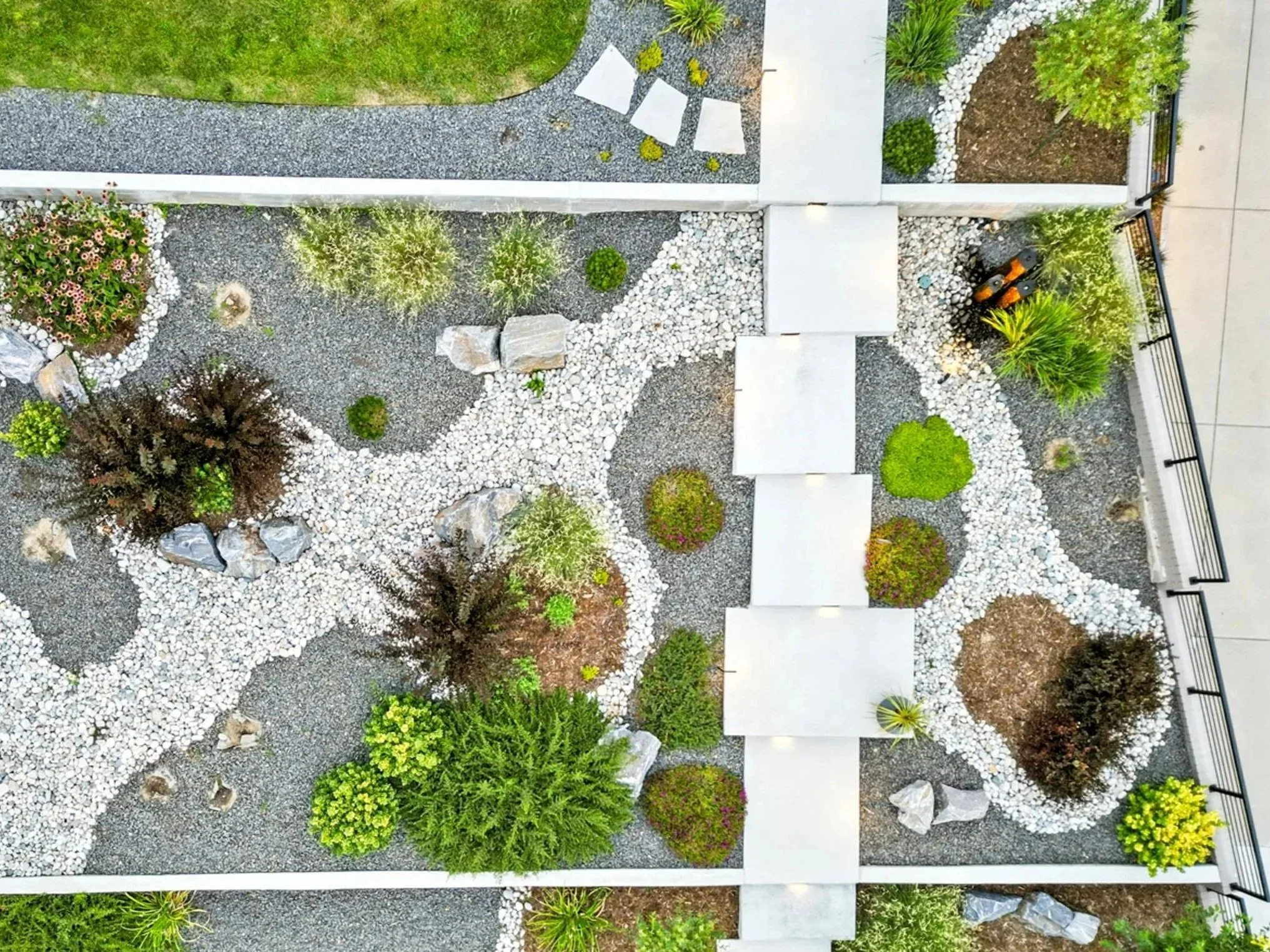 An aerial view of an outdoor landscaped garden with pathways, various plants, trees, rocks, and a patio area with benches.