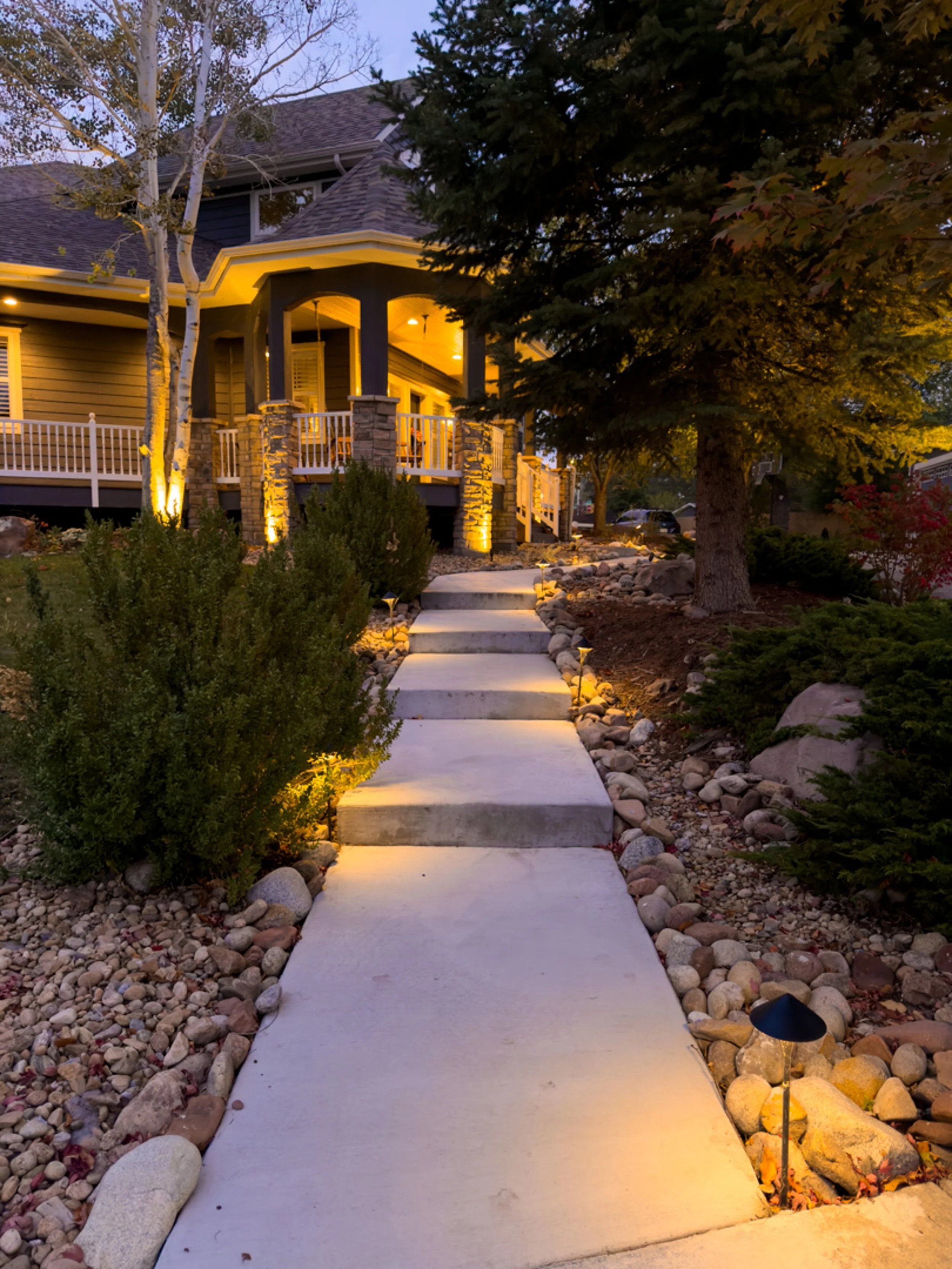 A well-lit front yard pathway with concrete steps, surrounded by rocks and plants, leading to a porch of a house at dusk.