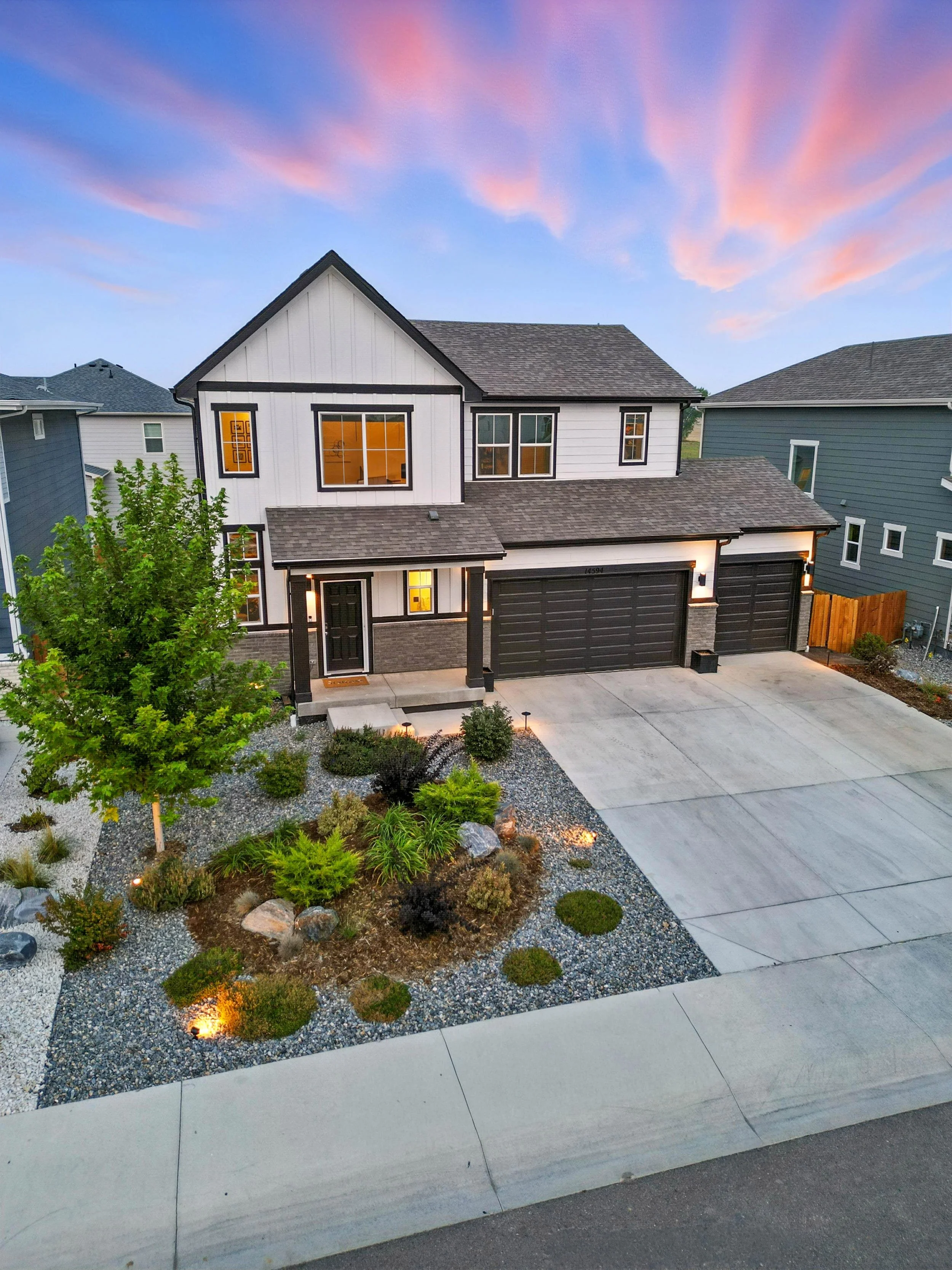 Modern two-story house with black garage doors, front porch, landscaped yard with trees, bushes, rocks, and pathway, during sunset with pink and purple sky.