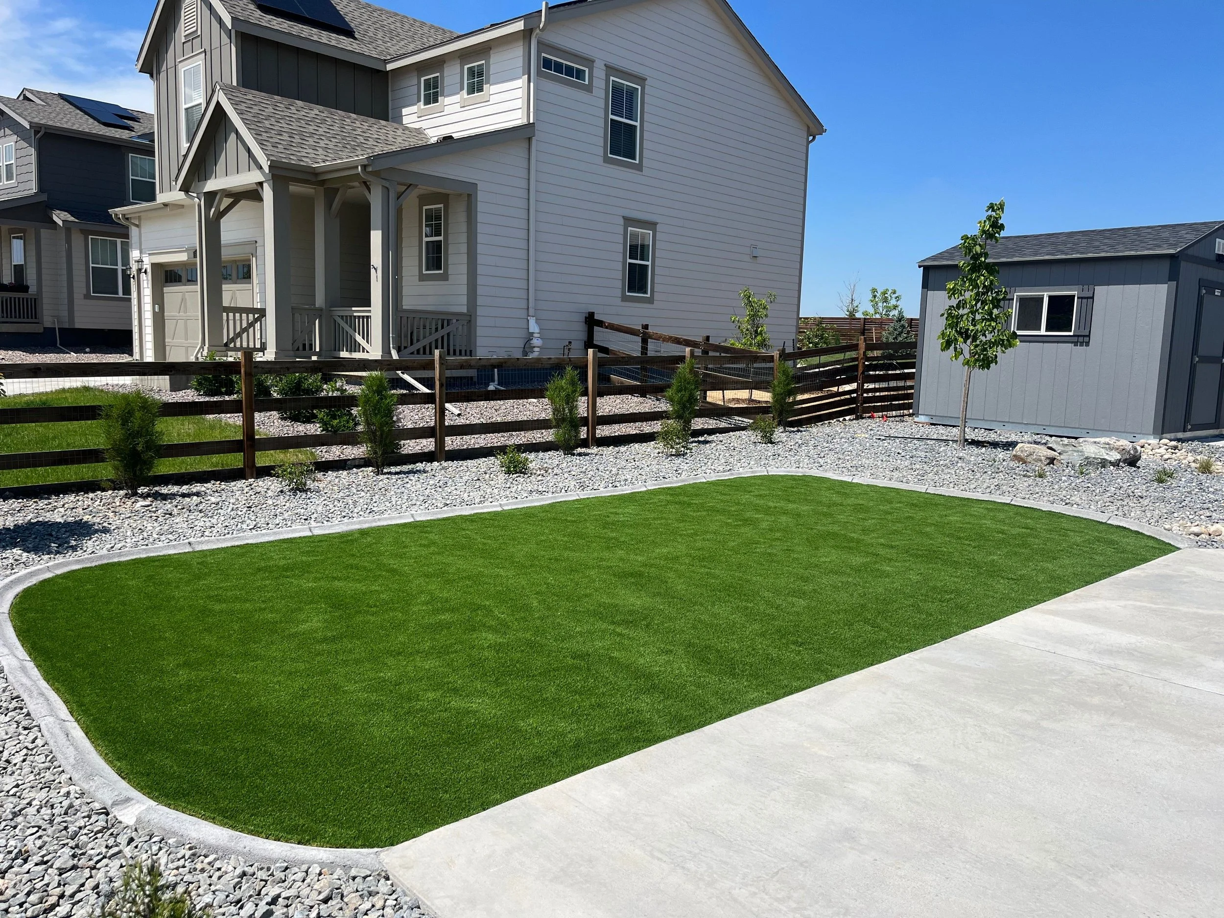 A backyard with a rectangular patch of artificial grass, a concrete patio, and a gravel border. There are small shrubs and a young tree with green leaves, a wooden fence, and outside houses in the background under a clear blue sky.