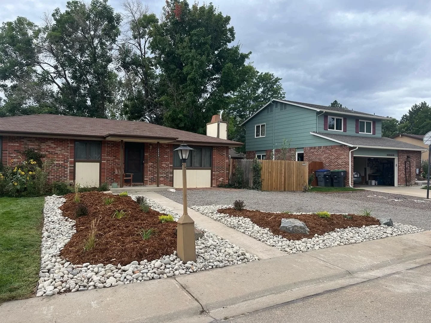 House with a brick exterior, front porch with a chair, landscaped yard with white rocks and mulched flower beds, gravel driveway, a street lamp, and neighboring houses in the background under a cloudy sky.