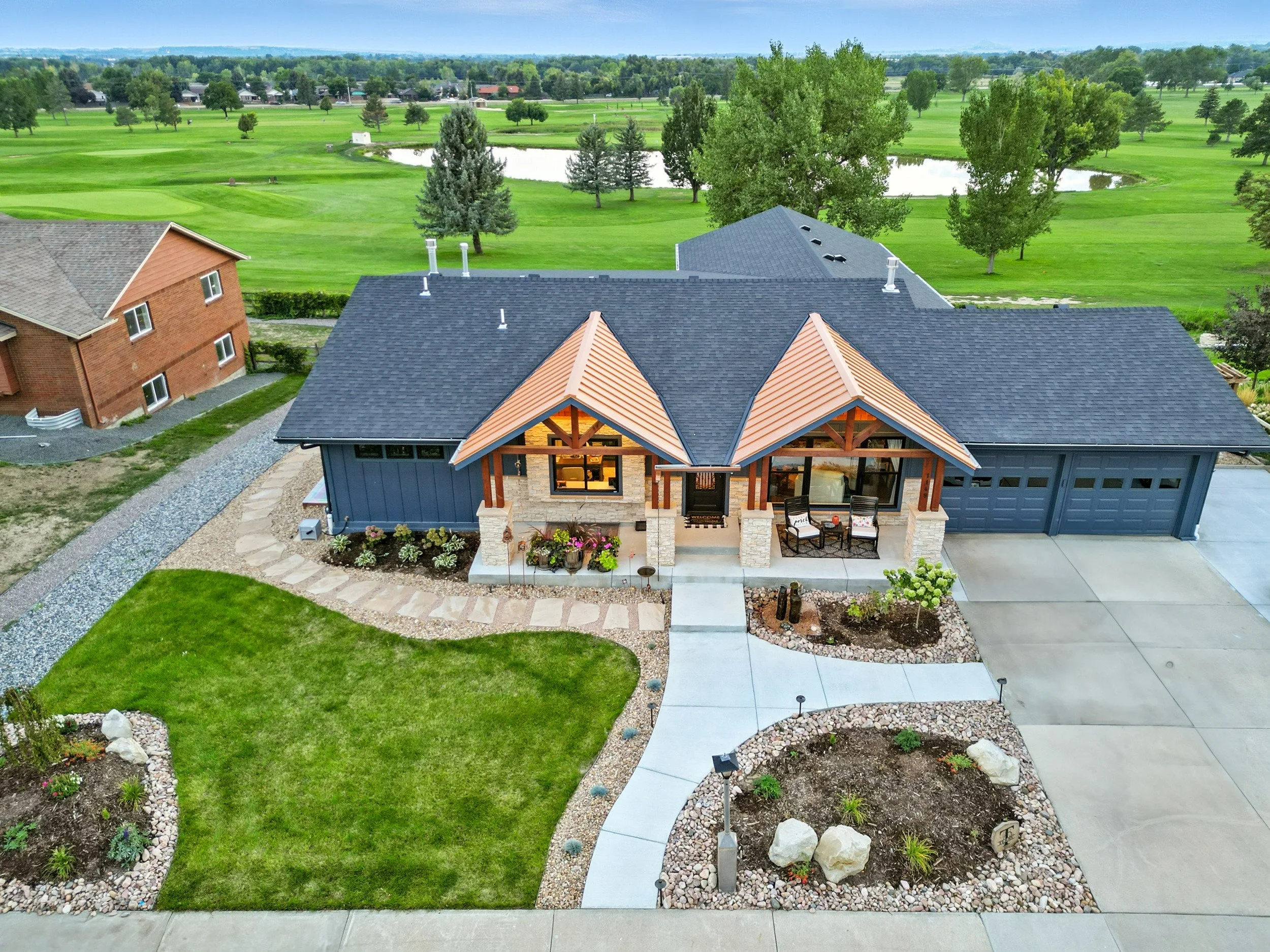 A modern house with a blue exterior, stone and wood accents, a dark gray roof, and a spacious driveway. The front yard has a small lawn, flower beds, and decorative rocks, while the background features a golf course with green grass, scattered trees,