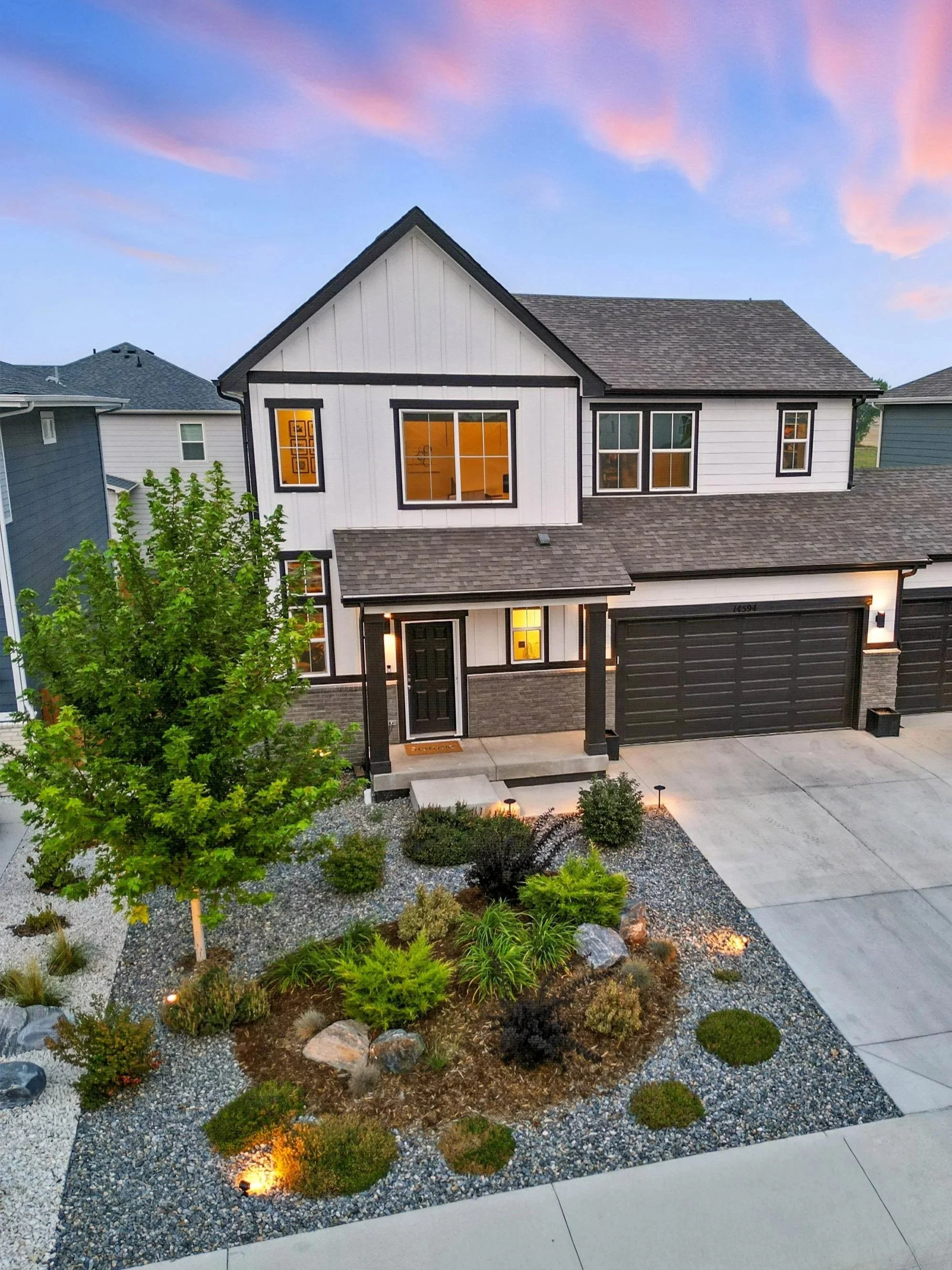 A two-story house with white siding, black trim, and a gray roof, illuminated by exterior lights, with a landscaped front yard featuring a tree, bushes, rocks, and decorative gravel, during dusk.