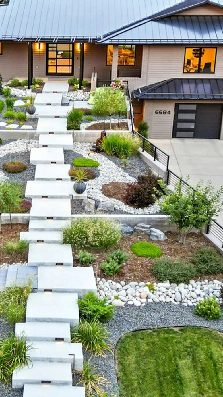 Front yard landscape with concrete walkways, plants, and decorative rocks leading to a modern house with large windows and a garage.