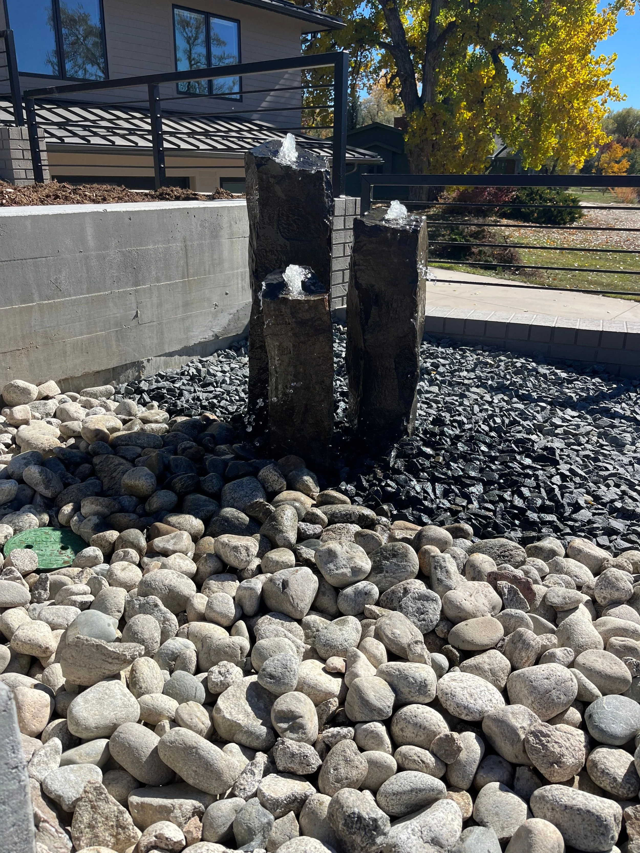 Outdoor decorative water feature with three black polished rocks with water flowing over them, surrounded by gray and white pebbles, with a modern house, trees with autumn foliage, and a sunny sky in the background.