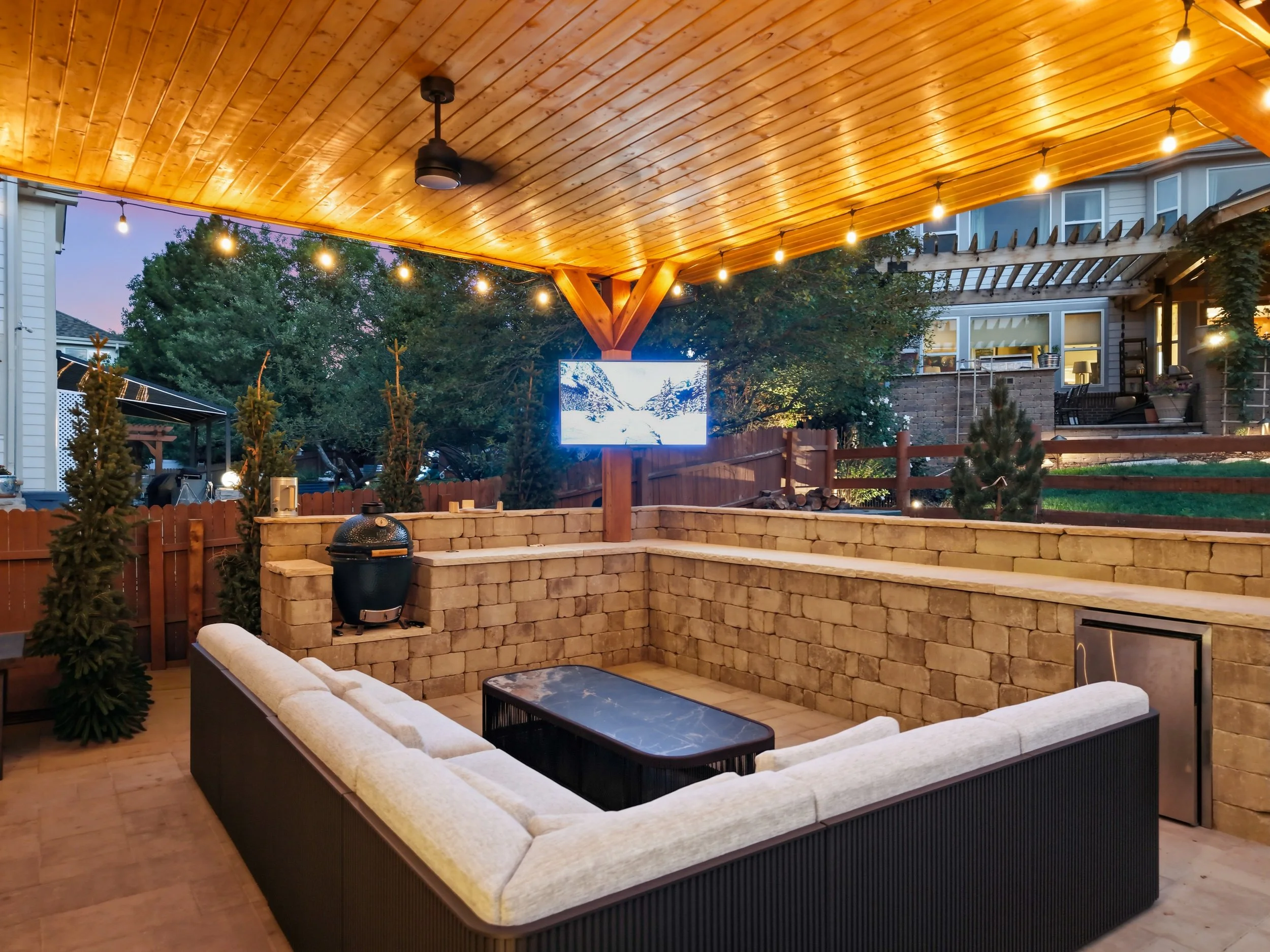 An outdoor patio with a wooden ceiling, string lights, a ceiling fan, and a built-in brick seating area with cushions. There are small trees and a flat-screen TV in the background.