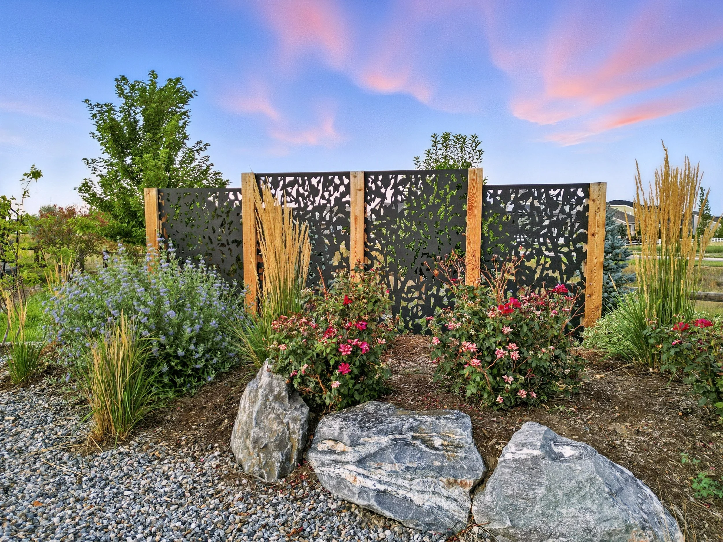 A garden with various plants and flowers, featuring a decorative black metal privacy screen with a nature-inspired cutout pattern, set against a sunset sky.