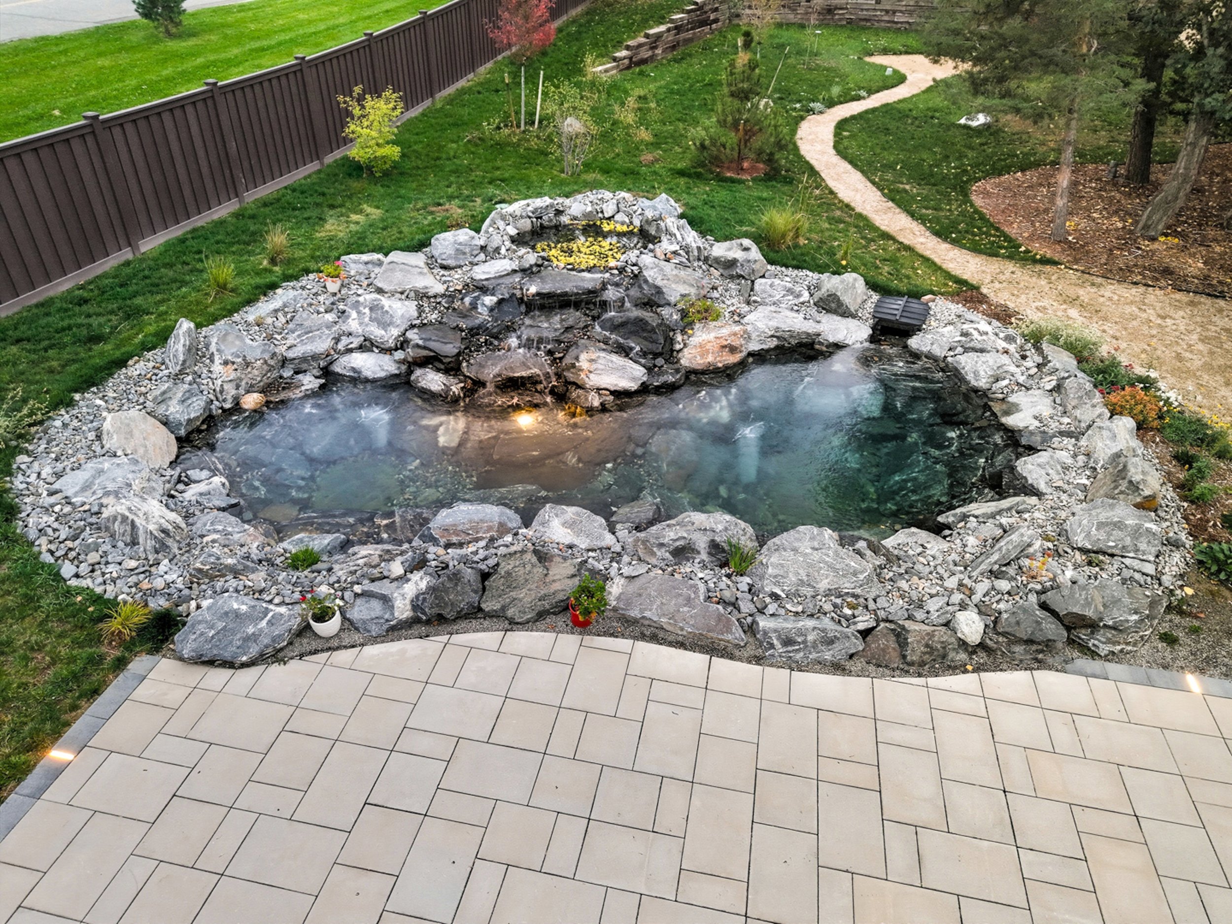 Backyard garden with a rock waterfall feature, surrounded by a gravel and tiled patio, with a winding pathway, trees, and a wooden fence.