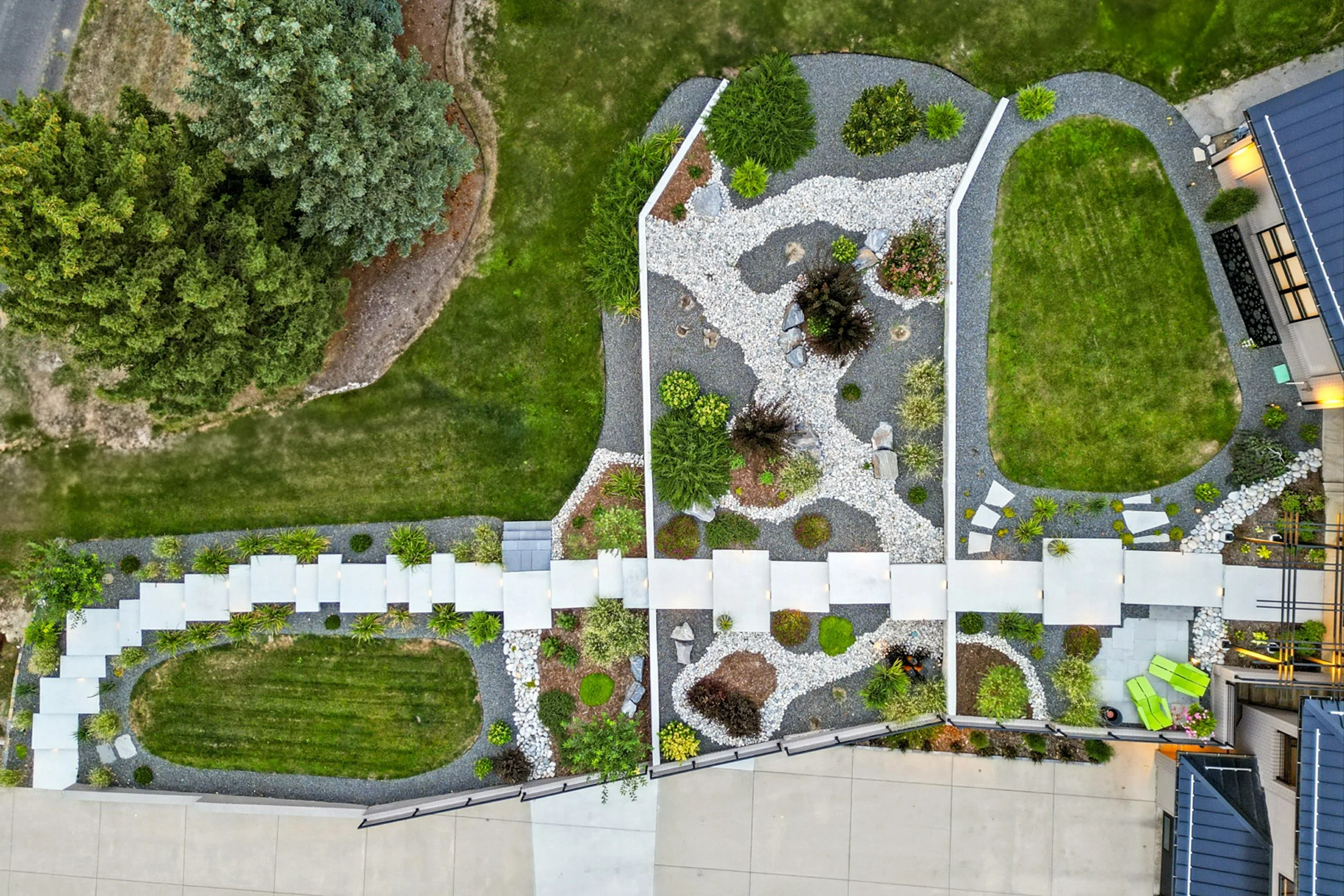 An aerial view of a landscaped garden with grassy areas, pathways, trees, shrubs, and decorative rocks surrounding a house with a small patio and bright green outdoor chairs.