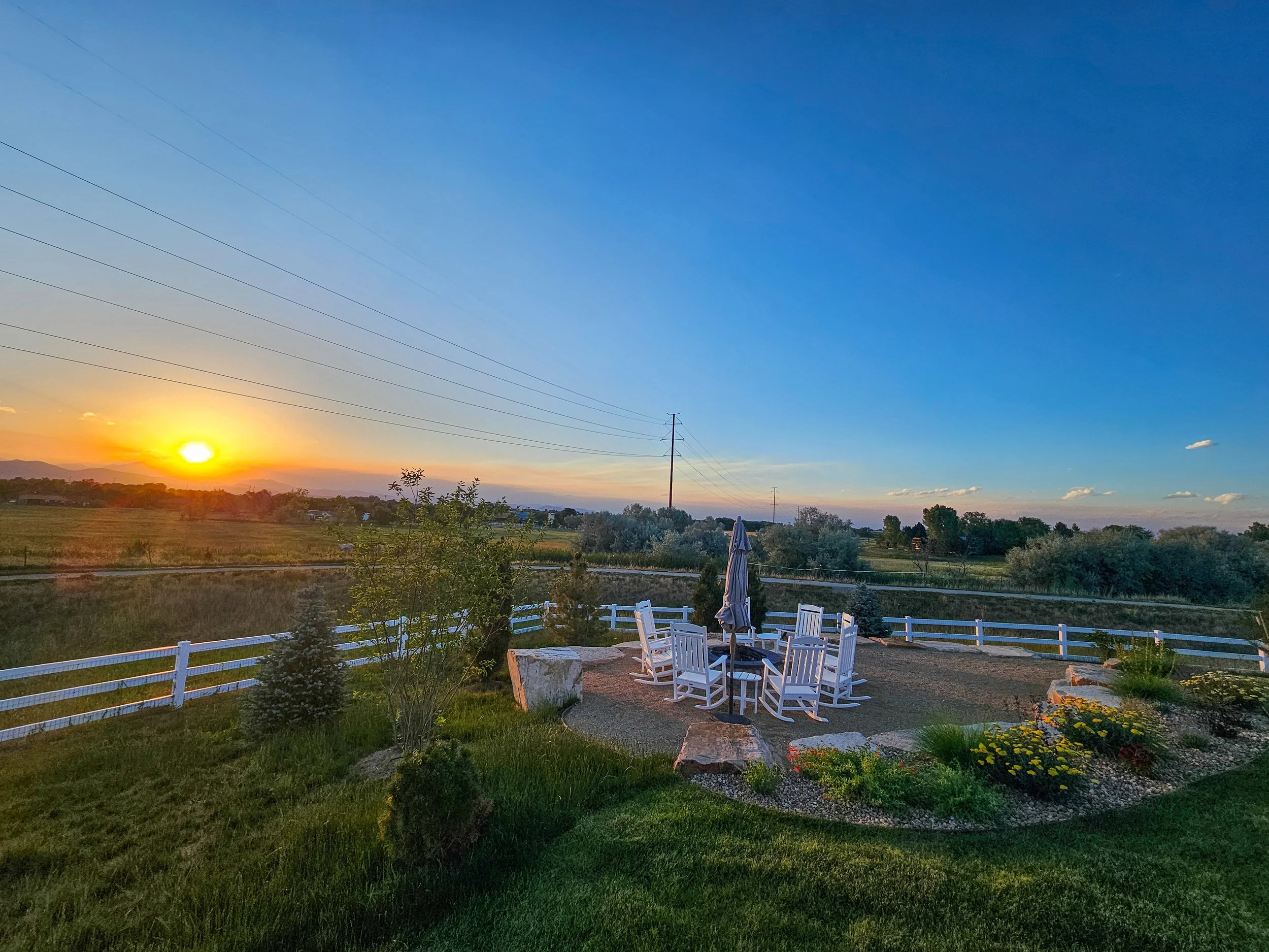 Sunset over a rural landscape with a white wooden fence, small trees, and a patio area with white chairs and a closed umbrella. Power lines run across the sky, which is mostly clear with some clouds.
